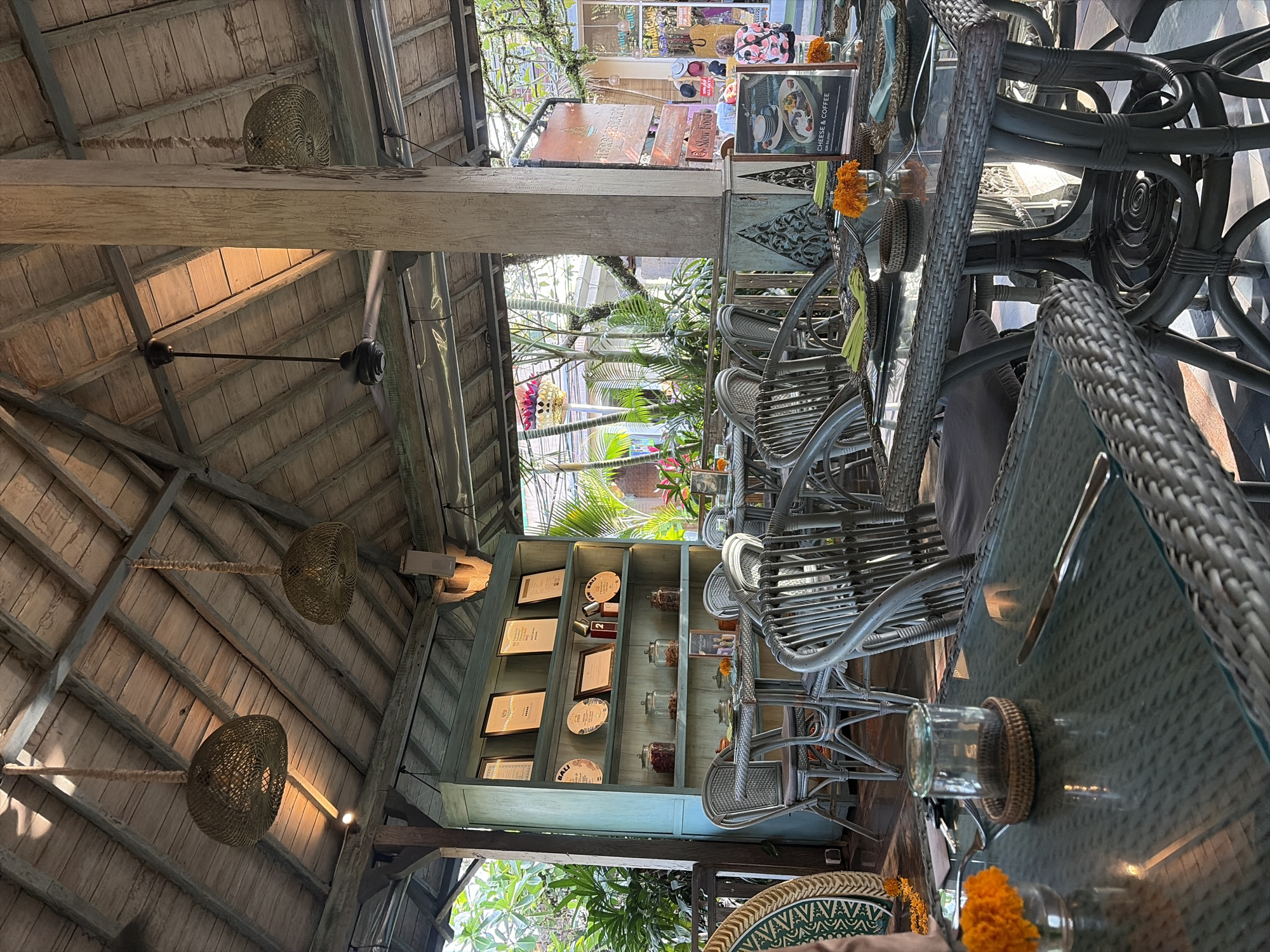 The image shows an outdoor dining area of a restaurant in Ubud, Indonesia. The area is covered by a thatched roof supported by wooden beams. Wicker tables and chairs are set for dining, with some tables featuring small arrangements of orange flowers. A light blue shelf unit filled with books, plates, and jars is visible against a wall. In the background, lush greenery and other buildings can be seen. A ceiling fan is mounted on the roof. The lighting suggests it is daytime, with sunlight filtering through the roof and trees. There are no people clearly visible interacting in the scene.