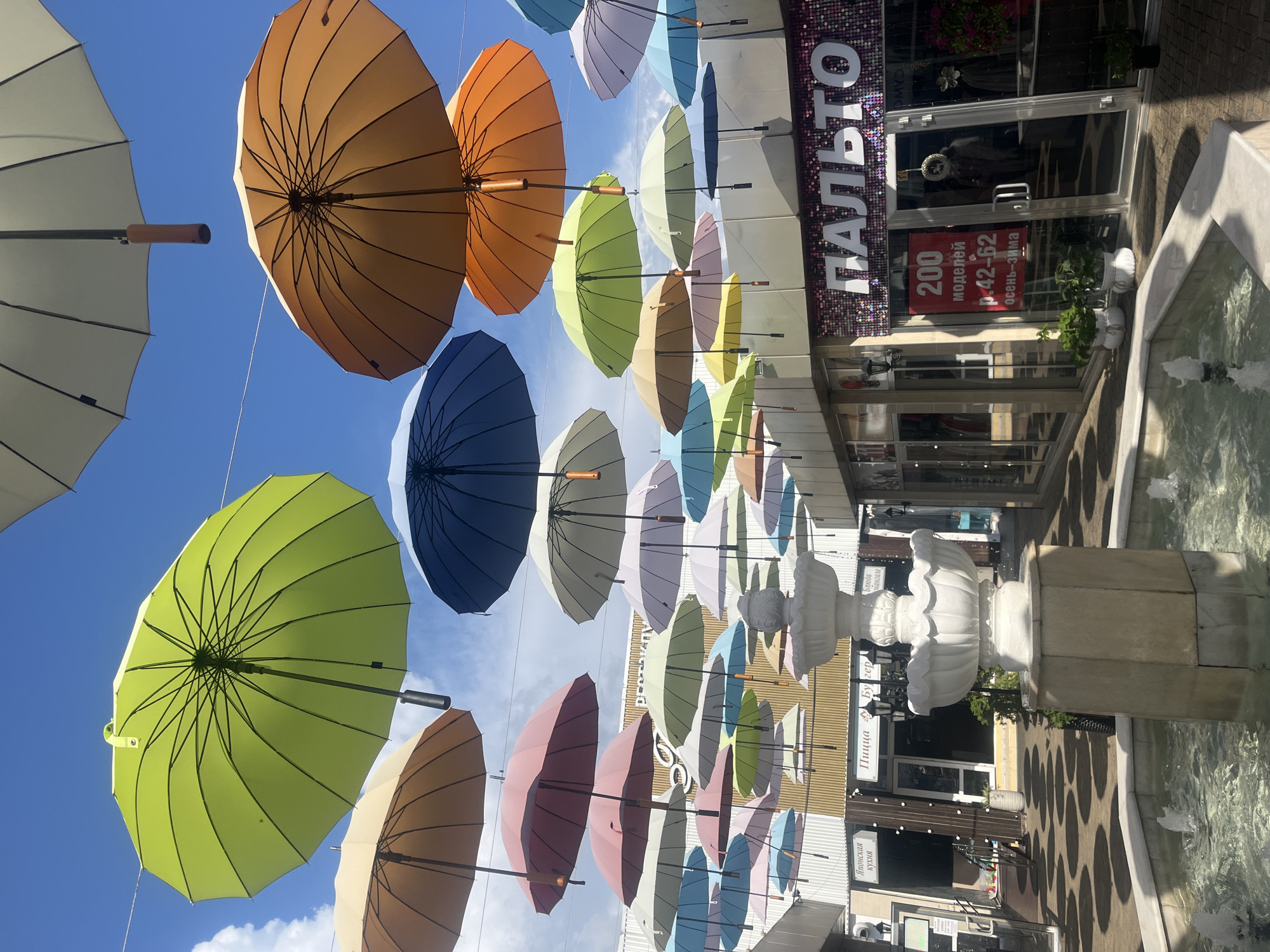 This image captures a vibrant outdoor scene, likely a pedestrian street or market, in Sochi, Russia. The focal point is a multitude of colorful umbrellas suspended in the air, creating a whimsical canopy against a bright blue sky dotted with white clouds. The umbrellas, in shades of green, orange, pink, blue, and yellow, are a dominant visual element, casting dappled shadows on the pavement below.
In the foreground, a tiered white fountain with spouting water adds a touch of elegance and refreshment to the scene. The fountain sits within a shallow basin, its surface reflecting the overhead umbrellas.
Behind the fountain and umbrellas, the facades of several shops are visible. One shop prominently displays a sign with Cyrillic lettering, "ПАЛЬТО" (Palto), meaning "coat," suggesting it is a clothing store. Another sign reads "200 моделей р. 42-62 осень-зима," indicating a sale of 200 coat models in sizes 42-62 for the autumn-winter season. Other shop signs with Russian text like "Японская кухня" (Japanese cuisine) and "Пицца Бургер" are also visible, indicating a commercial area with diverse offerings.
The architecture suggests a modern commercial complex with glass storefronts, metal siding, and decorative lighting. The ground is paved with interlocking stones, and potted plants add greenery to the environment. No people are clearly visible in the frame, though reflections in the shop windows suggest some movement within the stores. The overall impression is one of a lively and visually appealing public space designed to attract shoppers and visitors.