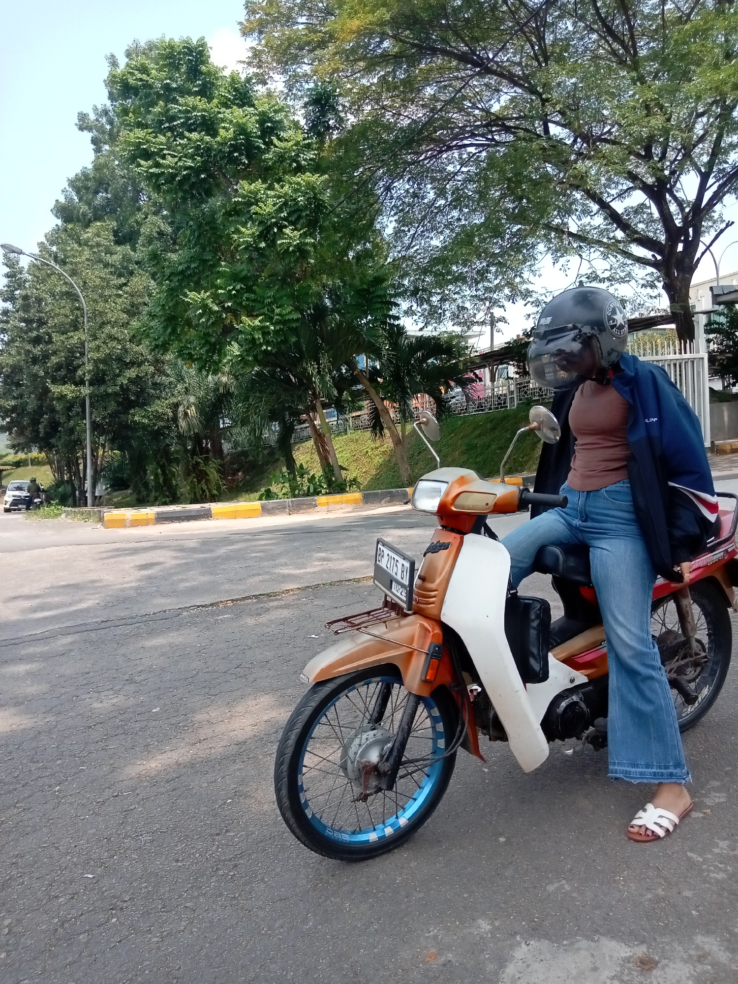 A young woman wearing a helmet and blue jeans is sitting on a scooter in a city street in Batam, Indonesia. She is wearing a brown shirt and a blue jacket. The scooter is orange and white. There are trees and a fence in the background. The weather is sunny.