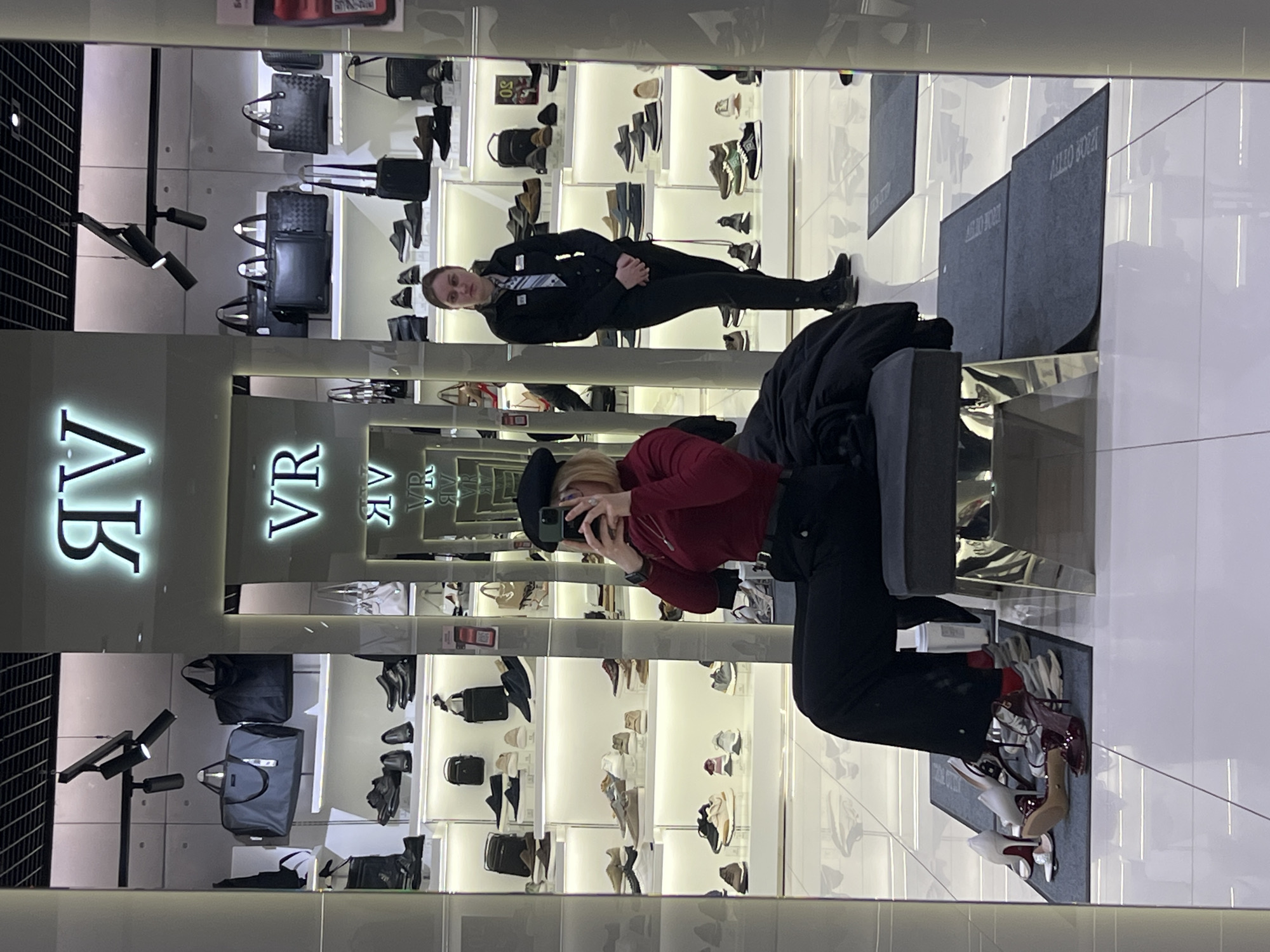 The image captures the interior of a modern, brightly lit shoe and accessories store, likely in Odesa, Ukraine. The scene appears to be taken as a mirror selfie or reflection.
In the foreground, a woman wearing a black beret, a red long-sleeved top, and black pants is seated on a grey armchair, holding a smartphone up to take a picture. In front of her, on a grey rug, several pairs of high-heeled shoes are displayed.
The store features extensive illuminated display shelves lining the walls, showcasing a wide assortment of footwear, including sneakers, boots, and dress shoes. Alongside the shoes, various handbags and duffle bags are also on display. The floor is a reflective, glossy white, mirroring parts of the store's interior and displays. Modern black track lighting fixtures are visible on the ceiling.
Further back in the store, a man, presumably a store employee, stands facing towards the camera. He is dressed in a dark jacket, dark pants, and a light-colored tie, with an ID badge visible on his chest.
Prominent illuminated signage with the letters "VR" is visible on a wall. On the grey floor rugs, the text "ALLO ZOLAN" can be partially seen, both directly and as a reflection. The store presents a clean, organized, and contemporary retail environment.