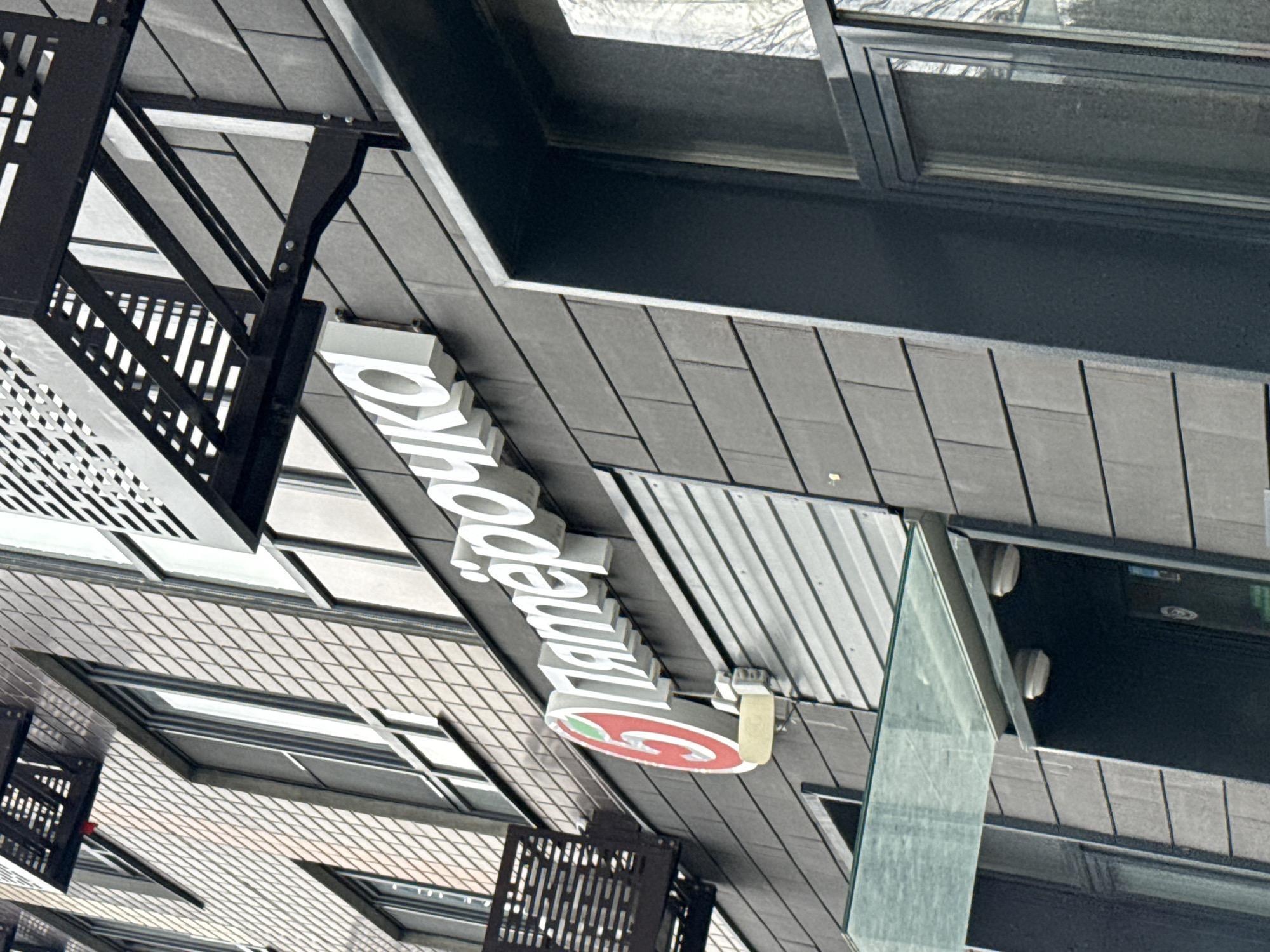 The image shows the exterior of a Pyaterochka supermarket, a Russian chain, located in Lyubertsy, Russia. The store features a prominent white sign with the Pyaterochka logo, which is a red circle with a white "5" inside, to the left of the text. The building's facade is made of dark gray, textured tiles. Above the entrance, a glass awning is installed. Metal fire escapes are visible on the upper floors of the building. The lighting suggests it is daytime, possibly overcast.
