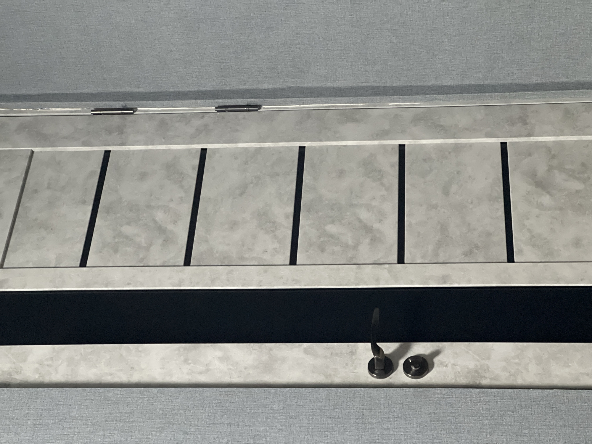 A close-up shot depicts a gray and white cabinet with black vertical accents. Two metal knobs are visible on the lower section of the cabinet. The surrounding environment is composed of gray patterned material. The location is not specified in the image.