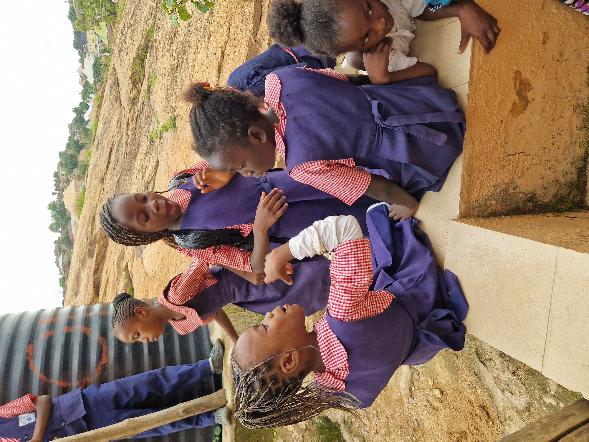 A group of five young girls in matching purple school uniforms with red and white checkered collars and cuffs are gathered outdoors on a rocky, uneven terrain. The girl in the foreground on the left is sitting and looking to her left, with her right arm bent and hand extended, as if speaking. Her hair is styled in thin braids. Behind her, a girl in a similar uniform is standing and looking forward with an open mouth, possibly speaking. She has her hair braided. To her right, another girl in the same uniform is also standing and looking away. In the far right, a girl is lying down and looking up with her chin resting on her hand, wearing a white top. Another girl is partially visible in the background, also in a school uniform. A large, ribbed, dark cylindrical object, possibly a water tank, stands to the left of the frame. The environment appears to be a hillside with scattered buildings and vegetation in the distance. The ground is composed of dirt and rocks. The lighting suggests it is daytime.
