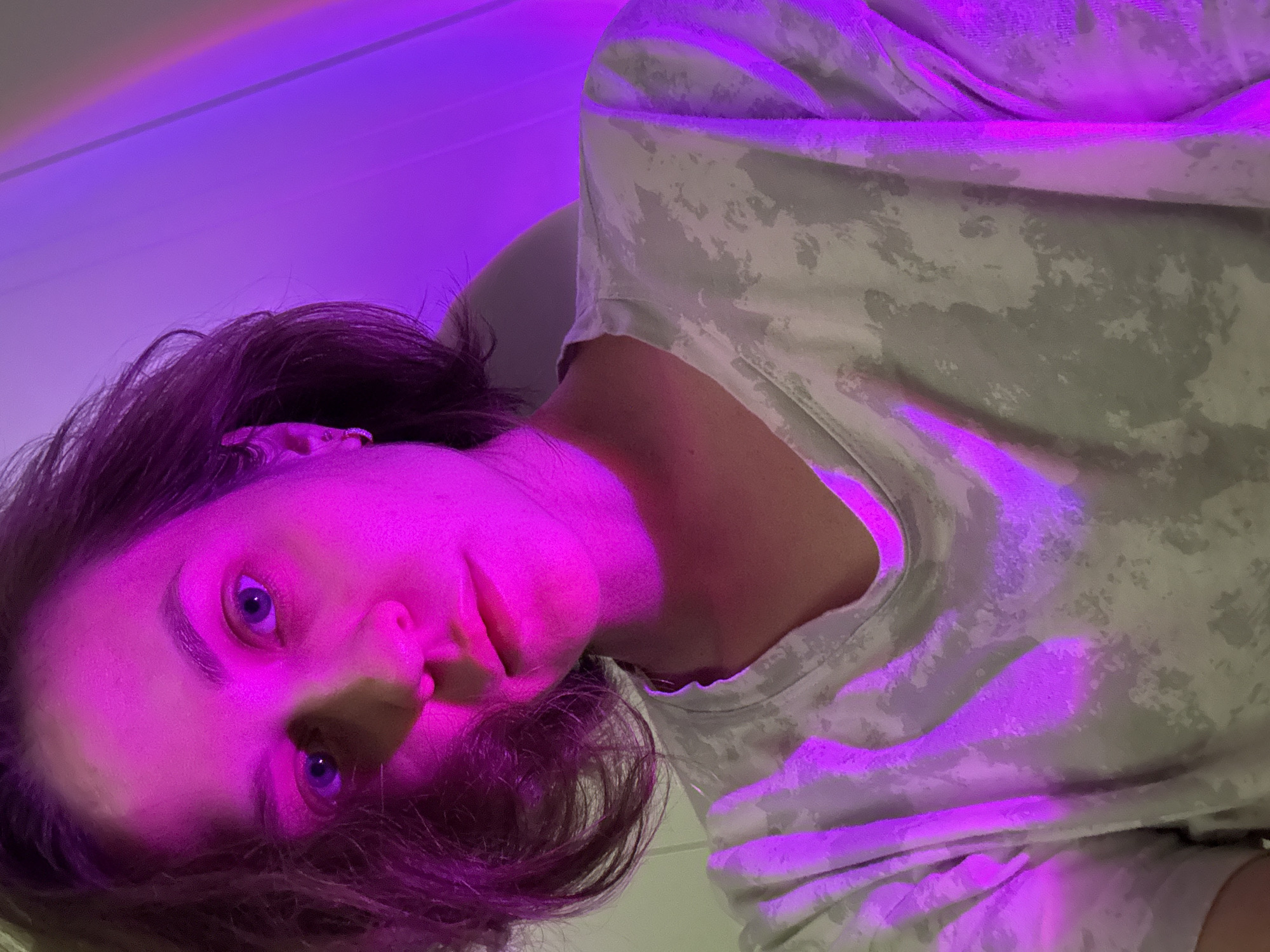 The image is a close-up, horizontally oriented portrait of an individual, likely an adult female, looking directly at the camera. The subject has dark, medium-length hair and blue eyes, and her face, hair, and upper body are intensely illuminated by a dominant magenta and purple light source, suggesting an indoor setting with mood lighting. Her skin and features are deeply colored by the strong light.
She is wearing a light-colored, short-sleeved t-shirt with a subtle, muted camouflage or abstract pattern, which also reflects the vibrant purple hues. The perspective is from a low angle, looking upwards at the person's face, giving the impression that the individual might be lying down or the camera is held at an unconventional angle.
The background is largely indistinct, showing gradients of the same purple light across what appears to be a wall or ceiling. There are no specific objects or identifying features in the background. The person holds a neutral to slightly contemplative expression, with her eyes wide open and focused on the viewer. No specific activity is taking place other than posing for the photograph. There is no visible text. The provided location context of Pervomaysky, Russia, cannot be discerned from the image itself.