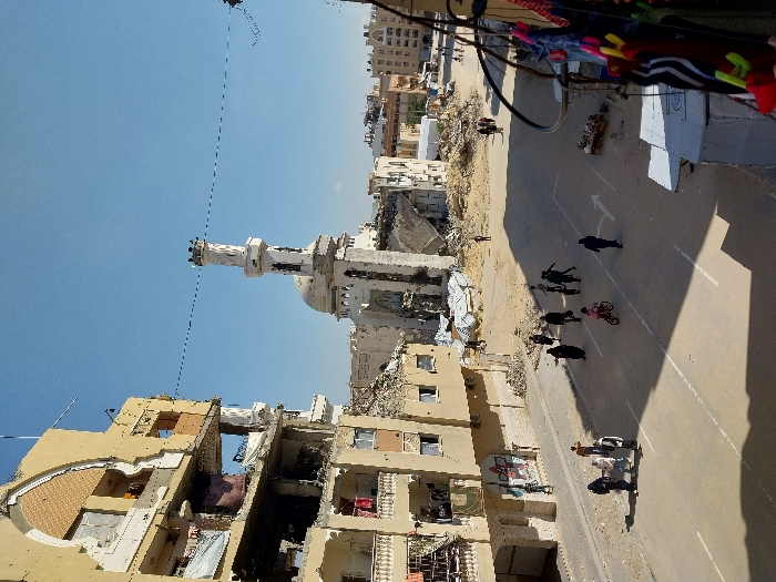 The image presents an urban scene in Khan Yunis, Palestinian Territory, characterized by significant structural damage under a clear blue sky. On the left, a multi-story, tan-colored building displays extensive damage, with multiple floors having external walls blown out, exposing internal rooms, and damaged balconies from which clothing is visible. Debris is accumulated at its base.
In the midground, a paved street runs diagonally, marked with white lines and a right-pointing arrow. Approximately 15 individuals are present on the street; a group of 5-6 adults, predominantly women in dark clothing, is gathered on the left side, while others, including a child riding a pink bicycle, walk towards the right. A small cart with items is visible on the street's right edge.
In the background, a white mosque with a tall, slender minaret and a dome appears heavily damaged; substantial sections of its main structure and dome are collapsed, leaving large piles of rubble. Other distant urban buildings are visible, some also showing signs of damage. Extensive piles of debris, including concrete fragments and sand, are scattered across the scene, particularly around the mosque and along the street's edges. A single thin cable stretches across the sky. The scene is illuminated by strong sunlight, casting defined shadows on the street. On the immediate right foreground, partially obscured, various garments hang from what appears to be a display rack.