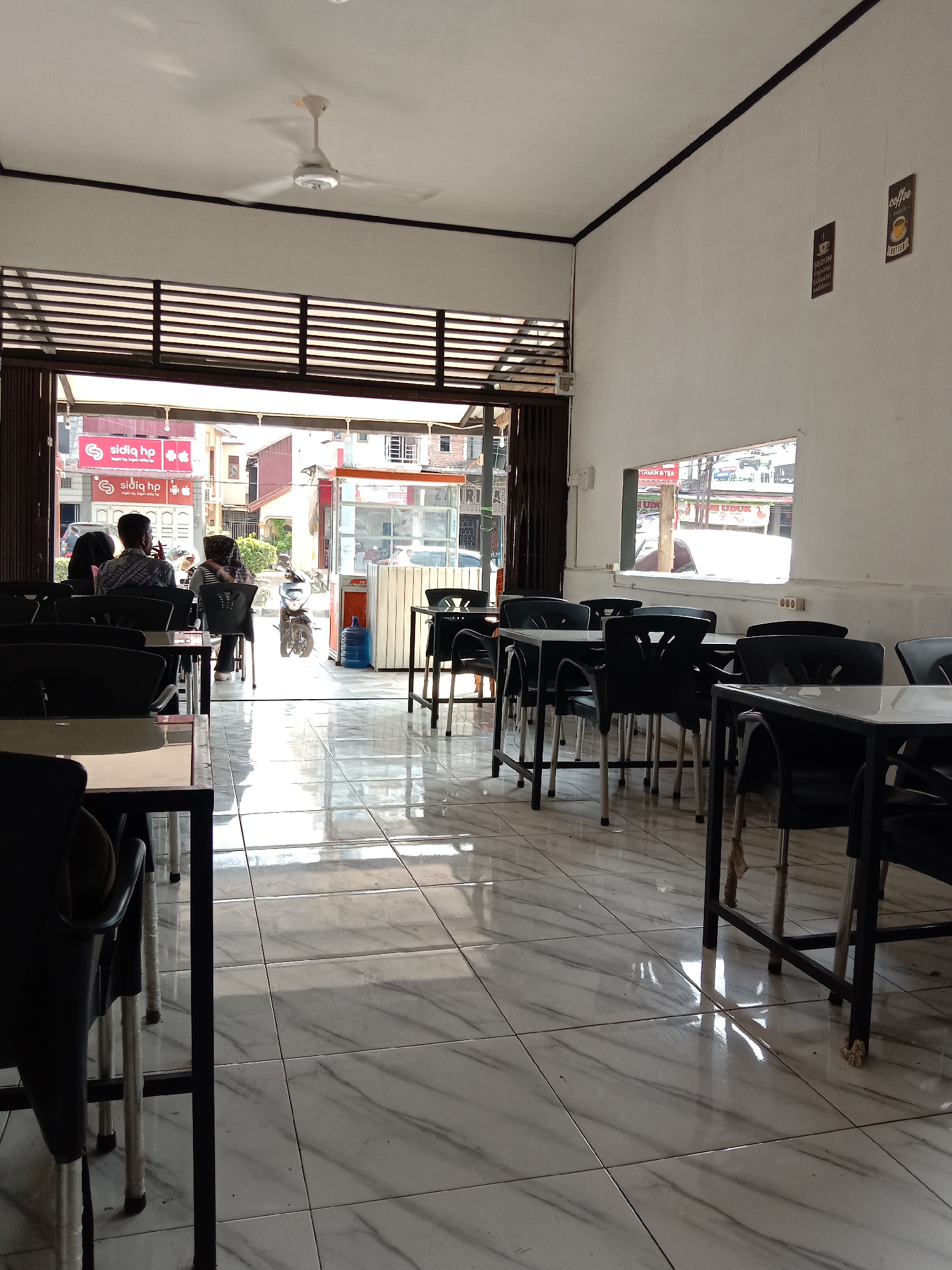 The image presents the interior of what appears to be a cafe or restaurant, likely in the city of Lhokseumawe, Indonesia, judging by the signage visible through the open entrance. Tables with black chairs are arranged across a tiled floor that reflects light from the exterior. The open entrance reveals a street scene with a vendor stall and several people, including a couple seated at one of the cafe's outdoor tables. The brand name "sidiq hp" is prominently displayed on a building across the street. Inside, the cafe has white walls adorned with coffee-related wall decor. A small, high window is also visible. A ceiling fan is centered in the room.