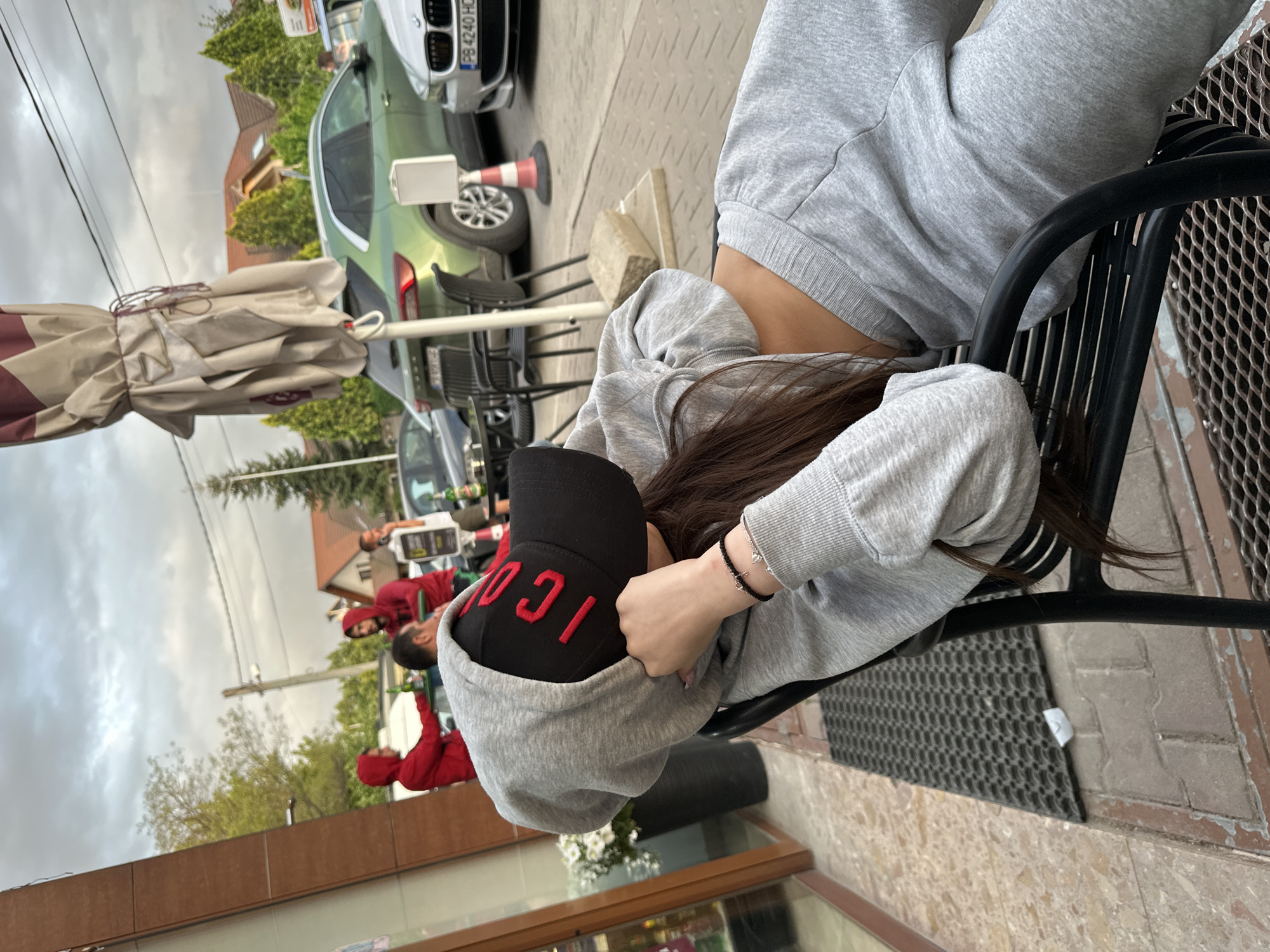 The image shows a person sitting on a black metal chair outdoors. The person wears a gray tracksuit and a black baseball cap with red lettering "ICON". Their face is obscured by the hood of the tracksuit and the cap. The person is holding their head in their hands.
In the background, other people wearing red clothes are visible, standing on the street. Cars are parked along the street. Buildings and trees are in the distance. There is also a white parasol. A green car can also be seen. The scene is located on a street with a pedestrian area next to a building. The setting suggests a cafe or restaurant area along a street.
The image doesn't show any clear indications of being located in Plovdiv, Bulgaria. The location requires further information.