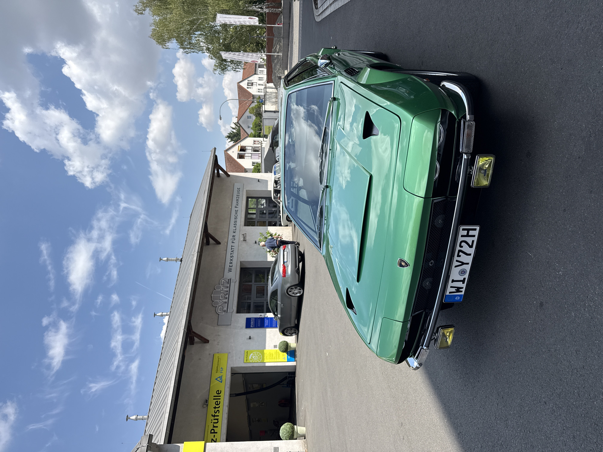 In Offenbach am Main, Germany, on a sunny day, a vibrant green vintage Lamborghini, identified by the license plate "WI Y72H," is parked on asphalt in front of a workshop labeled "PYRITZ Werkstatt für klassische Fahrzeuge." The workshop, with a grey, corrugated metal roof and white facade, has an open bay where a silver Audi is visible. A man in a blue shirt and grey pants is walking towards the Audi. The scene is framed by a bright blue sky with scattered white clouds and a line of green trees and houses in the background. The foreground shows the detailed front of the Lamborghini, including its distinctive headlights and hood vents.