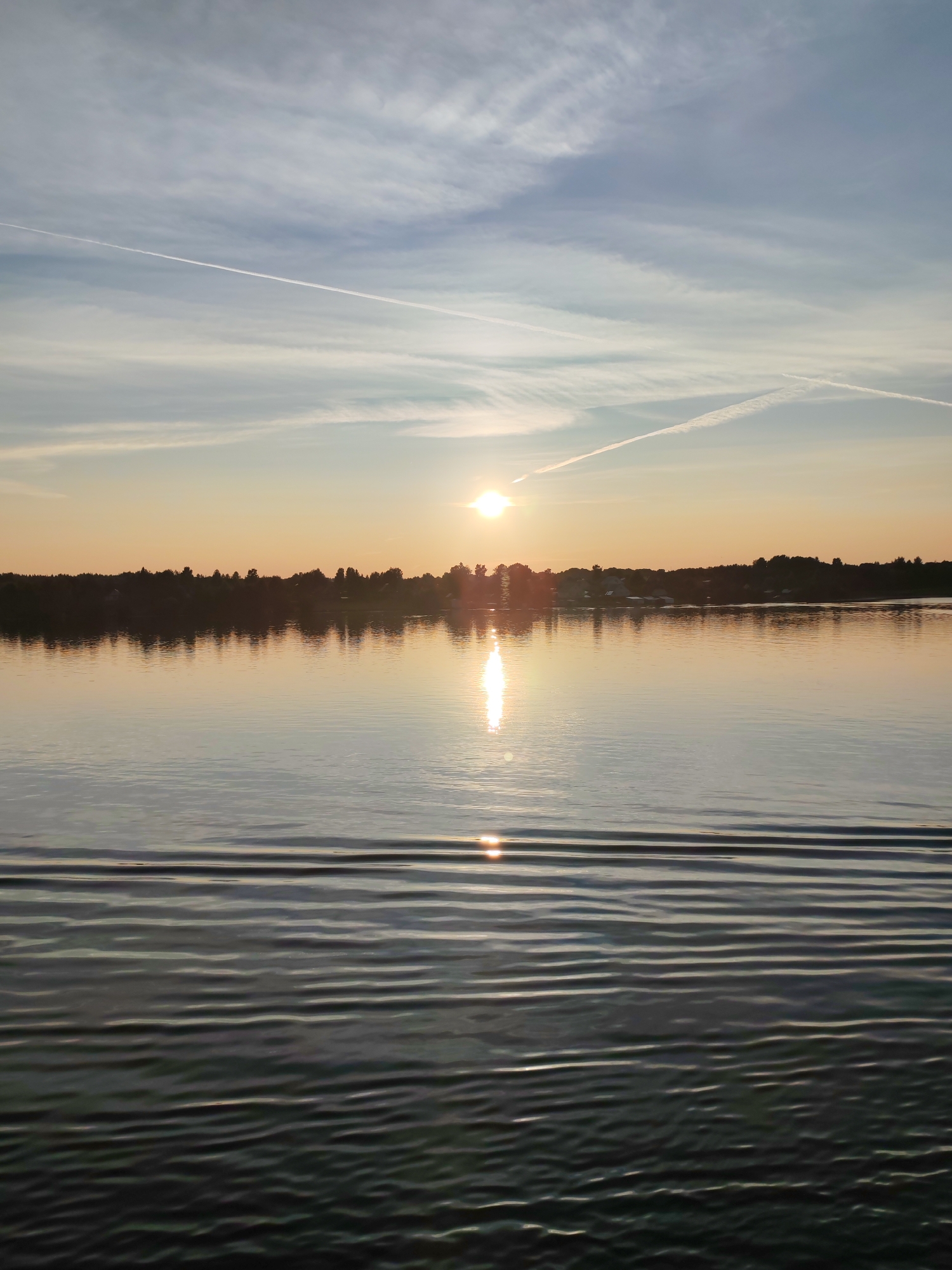 The image depicts a serene sunset over a calm body of water, likely a lake or wide river, in the vicinity of Bol'shoi Dvor, Russia. The sky is a gradient of soft oranges and blues, with wispy clouds and two distinct contrails from airplanes streaking across the upper portion. The sun, low on the horizon, casts a golden glow and reflects a shimmering path on the water's surface. A dark, silhouetted treeline dotted with small buildings marks the distant shore. The foreground of the water exhibits gentle ripples, suggesting a slight movement or disturbance from a vessel. No people are visible in the frame. The overall atmosphere is tranquil and picturesque, capturing the beauty of a natural landscape during twilight.