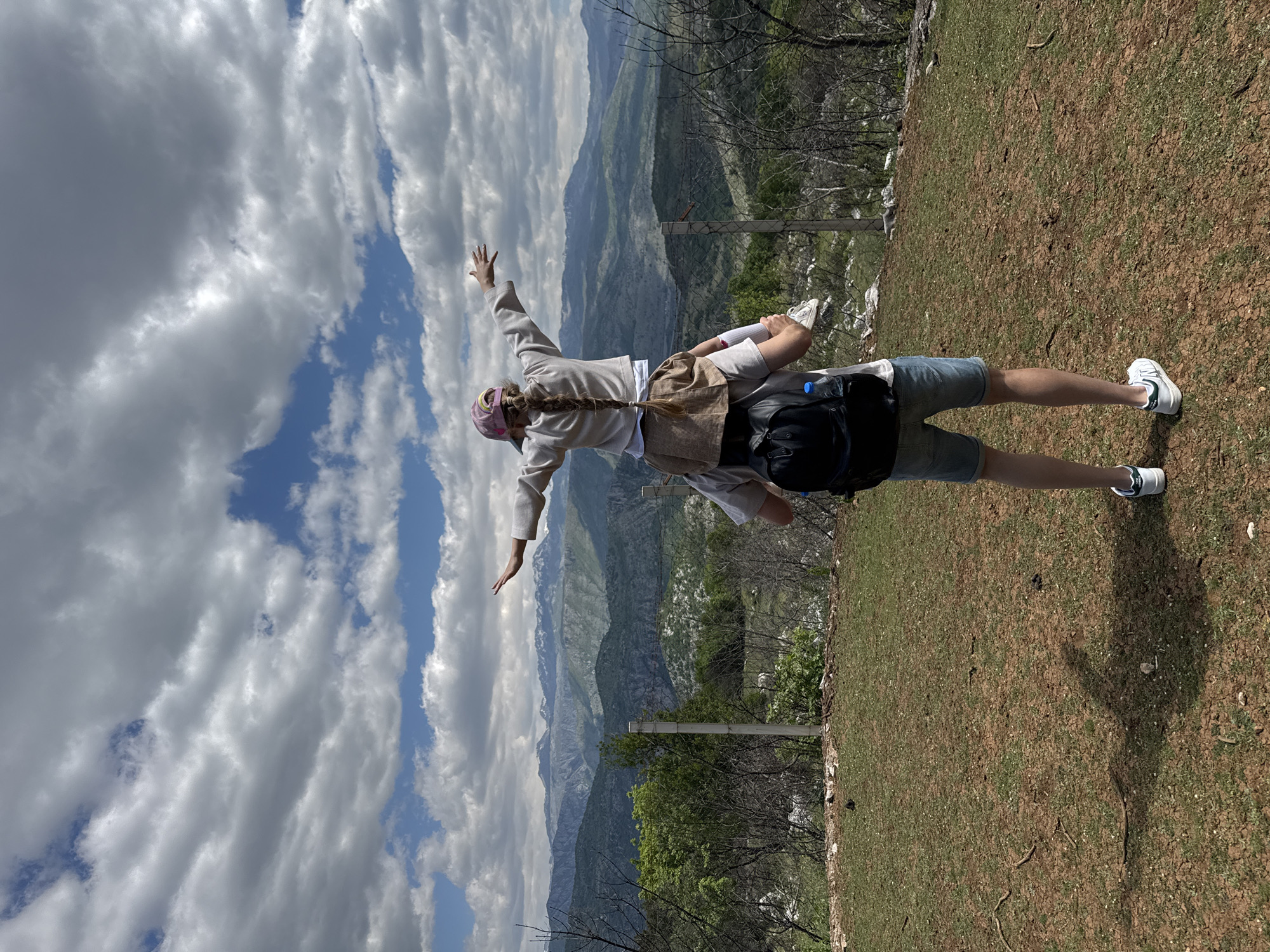 A man stands on a grassy and dirt-covered area, with a woman on his back appearing to be flying or mimicking an airplane. The location is Gornje Mrke, Montenegro, as indicated by the mountainous background and vegetation characteristic of the region. The man wears denim shorts and white sneakers, carrying a black backpack. The woman has a braid and wears a long-sleeved shirt, a light-colored skirt, and a pink hat or headband, with her arms extended outwards. In the background are mountains and a partly cloudy sky. A fence line and some sparse trees are visible.