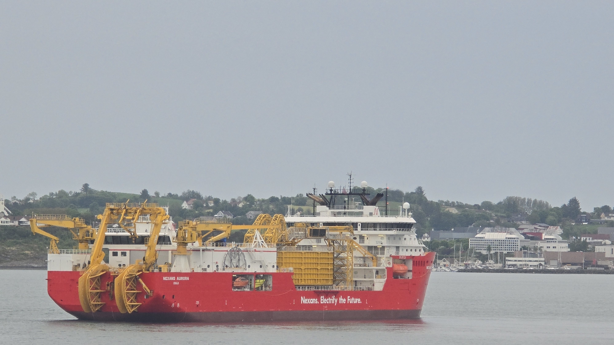 This image captures a large, modern industrial vessel, the "NEXANS AURORA," identified by its name on the stern, with "OSLO" below it. The ship features a distinctive red hull with a white superstructure and deck, and is equipped with prominent yellow specialized machinery, including crane-like structures and what appear to be cable-laying or handling systems on its aft deck. The slogan "Nexans. Electrify the Future." is clearly visible in white text on the port side of the hull.
The ship is situated on calm, grey water, suggesting it is in a bay, fjord, or harbor. In the background, a green, hilly coastline rises, dotted with various buildings. These include scattered houses, a white building with a steeple on the far left (possibly a church), and a larger, multi-story white building with numerous windows on the right side of the frame. Several smaller boats, possibly leisure craft, are moored near the shore on the right.
The sky is overcast and uniformly grey, indicating a cloudy daytime, likely with soft, diffused light. There are no visible people on the ship or along the distant shoreline. The scene conveys a sense of maritime industrial activity set against a tranquil, possibly Scandinavian, coastal landscape.