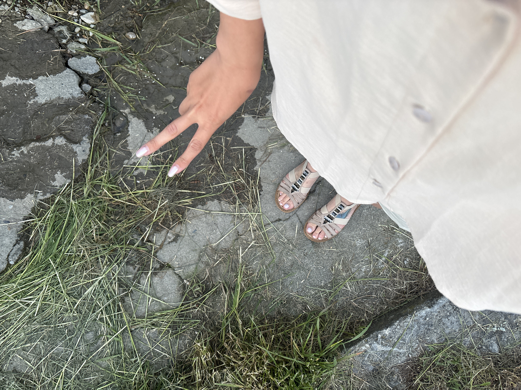 A top-down photograph shows a person standing outdoors on cracked concrete interspersed with patches of grass. The person wears a light-colored, button-down shirt or dress and tan sandals with beaded straps. Their left hand is raised, making a peace sign. The setting appears to be a semi-urban or edge-of-city location in Sochi, Russia. The ground consists of cracked concrete slabs with weeds and grass growing between them. There are no other people or objects visible in the photograph. The scene is static, with no apparent actions occurring.