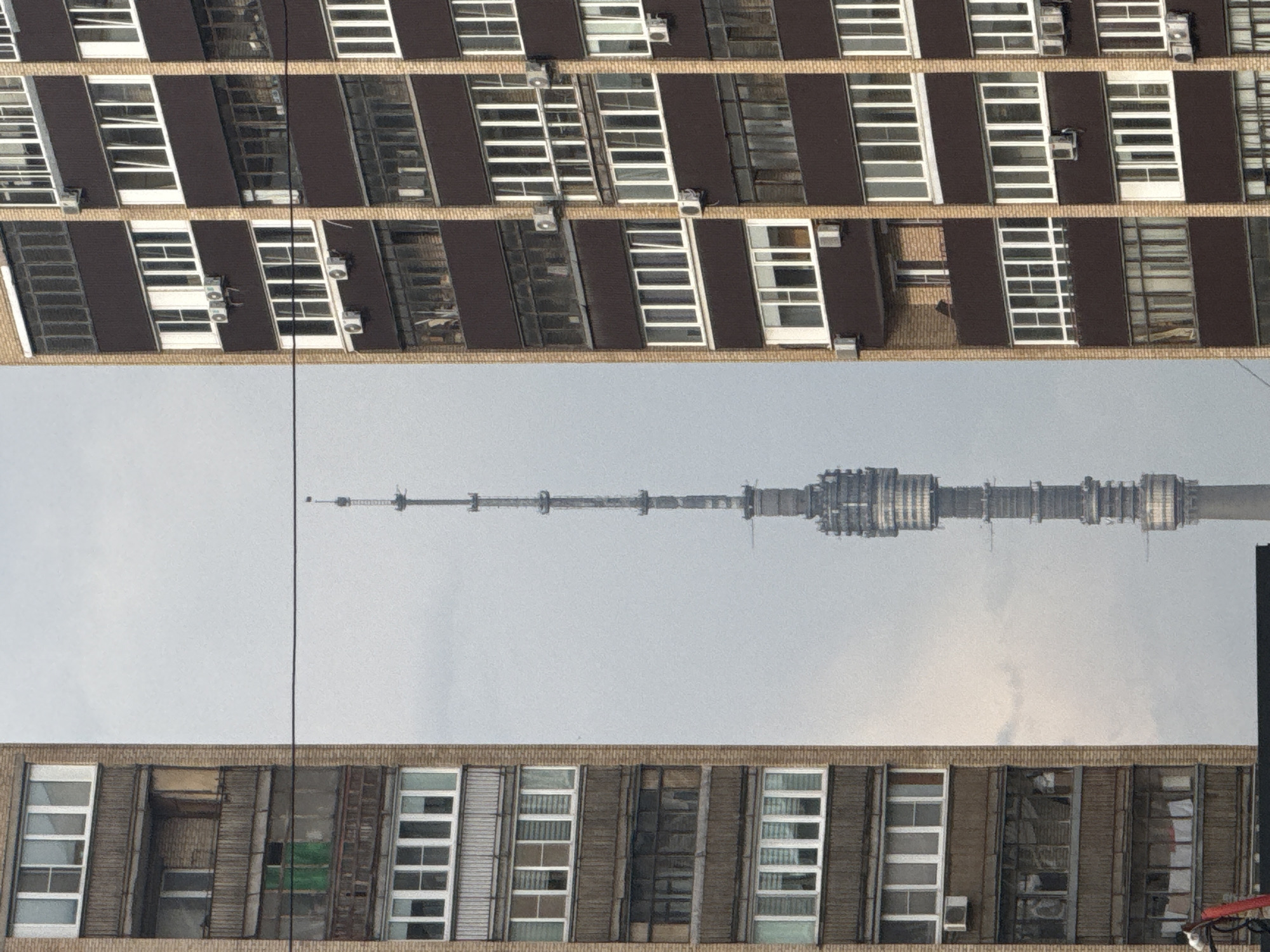 The image presents an upside-down view of Moscow's Ostankino Tower, positioned between two buildings. The buildings are of similar brick construction with rows of windows, some of which have air conditioning units. The sky is overcast. A thin, vertical wire is visible, possibly an electrical cable or a cable for a crane. There are no people visible in the scene.