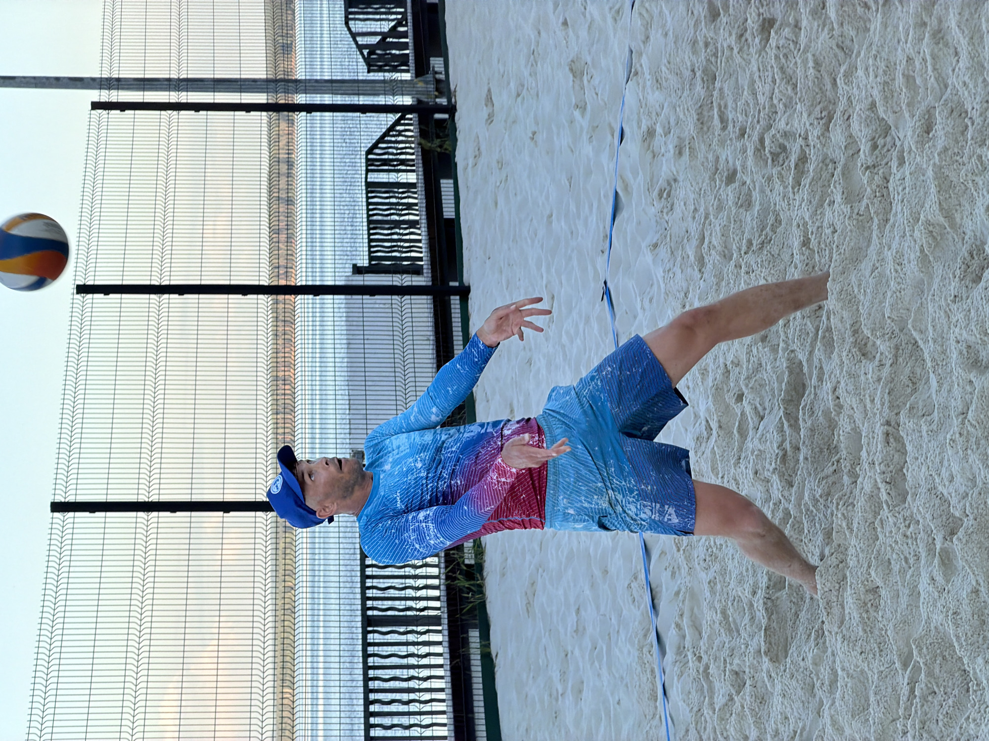 A bald Caucasian man wearing a blue baseball cap, a long-sleeved blue and red gradient shirt, and blue patterned shorts is playing beach volleyball. He is positioned on a sandy court with his arms extended, preparing to receive a volleyball that is in the air above and to his right. The background shows a metal fence, water, and some structures. The man's expression suggests concentration and focus on the game. The scene appears to be outdoors, possibly in a recreational area.