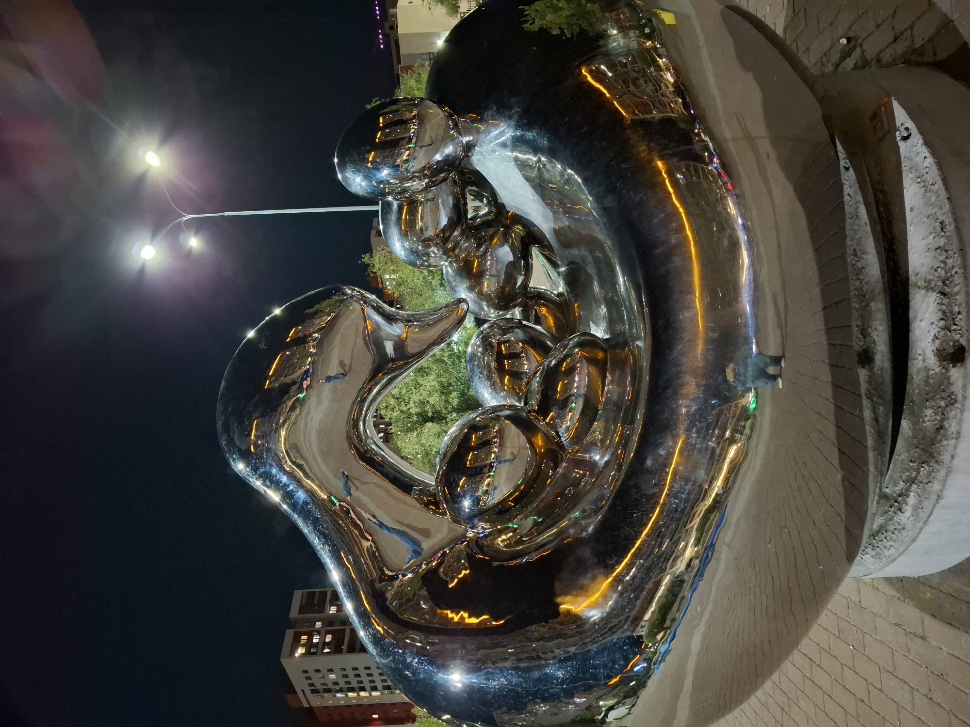 An outdoor, night-time, low-angle shot captures a large, highly reflective, chrome sculpture in Tyumen, Russia. The sculpture, with its smooth, undulating surfaces and spherical elements, mirrors the surrounding environment. Streetlights with multiple bulbs illuminate the scene, casting a bright glow. In the background, multi-story buildings with illuminated windows are visible, suggesting an urban setting. Trees with green foliage are interspersed between the buildings and the sculpture. The ground is paved with interlocking bricks. A lone individual, dressed in dark clothing, is reflected in the sculpture, standing near its base. The sculpture itself appears to be situated on a concrete pedestal. The overall atmosphere is one of modern urban art and nighttime ambiance.