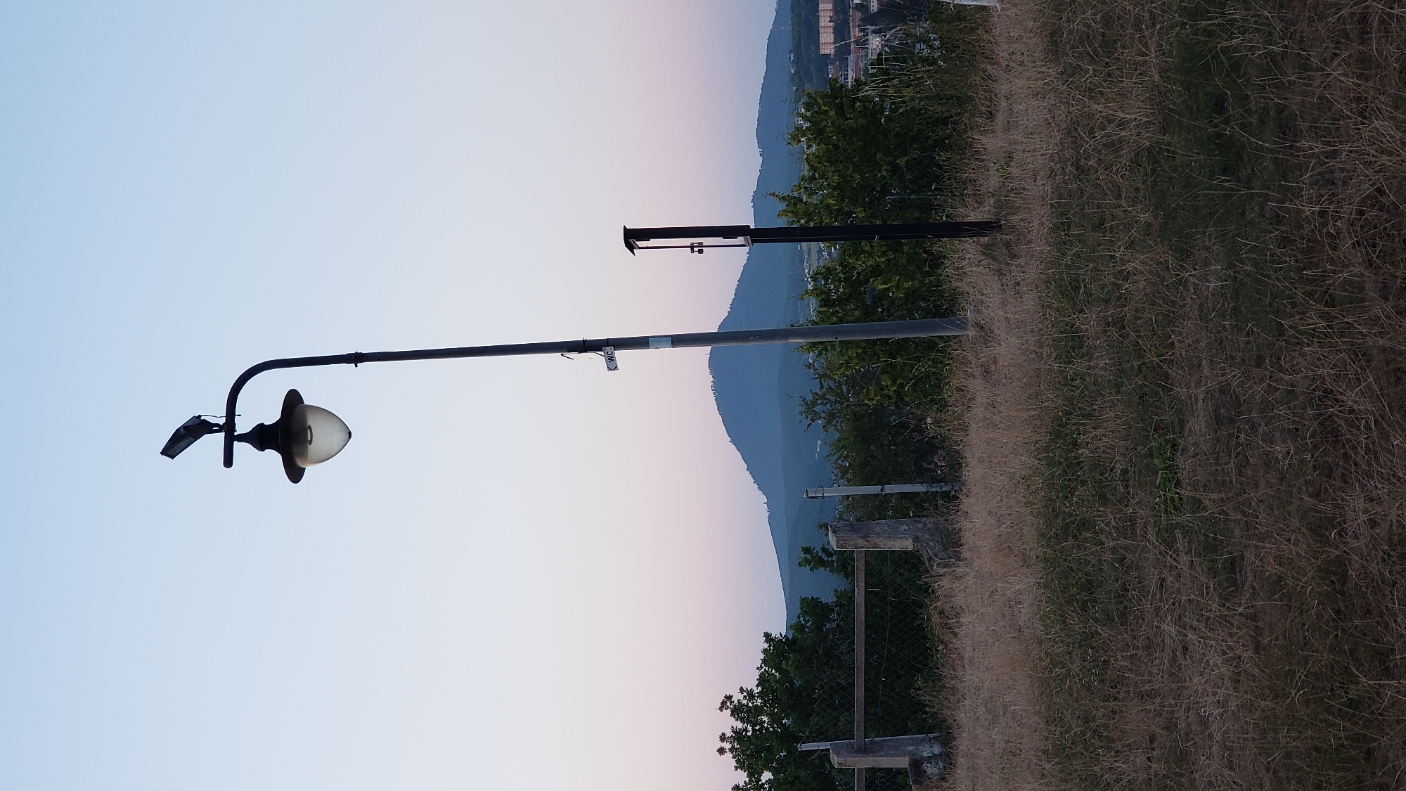 The image depicts an outdoor scene, likely at dusk given the soft pastel colors in the sky. In the foreground, dry, overgrown grass covers the ground. A tall, dark lamppost with a solar panel on top and a decorative lantern is a prominent feature in the middle ground. To the right of the lamppost, another dark, rectangular object, possibly a signpost or electrical box, stands tall. Behind the lamppost, a chain-link fence with concrete posts runs horizontally, separating the foreground from the background. Beyond the fence, lush green trees and bushes are visible. In the distance, a large, rounded mountain dominates the landscape, silhouetted against the fading light. The sky transitions from a pale blue at the top to soft pinks and purples near the horizon. The presence of buildings on the right side of the frame suggests an urban or suburban setting. Without additional context, the specific location within Prešov, Slovakia cannot be determined from the image alone, but the overall scene suggests a park or elevated viewpoint overlooking the city and surrounding natural landscape.