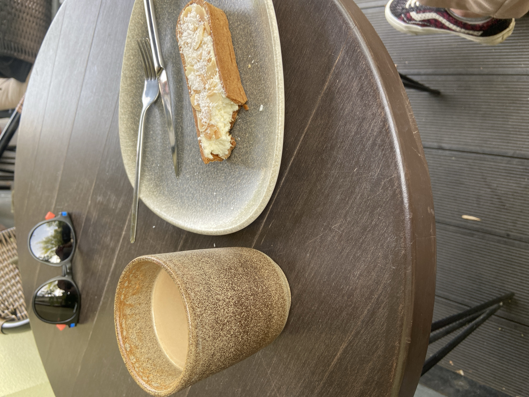 An outdoor cafe scene shows a circular brown wooden table on a grey wooden deck. On the table are a speckled brown ceramic mug filled with a light brown beverage, a grey ceramic plate with a slice of almond tart and a fork and knife, and a pair of black sunglasses with colorful accents. A pair of feet wearing a sneaker with red and black accents and tan pants are visible at the bottom right of the image. The background shows a wicker chair and a hint of green foliage.