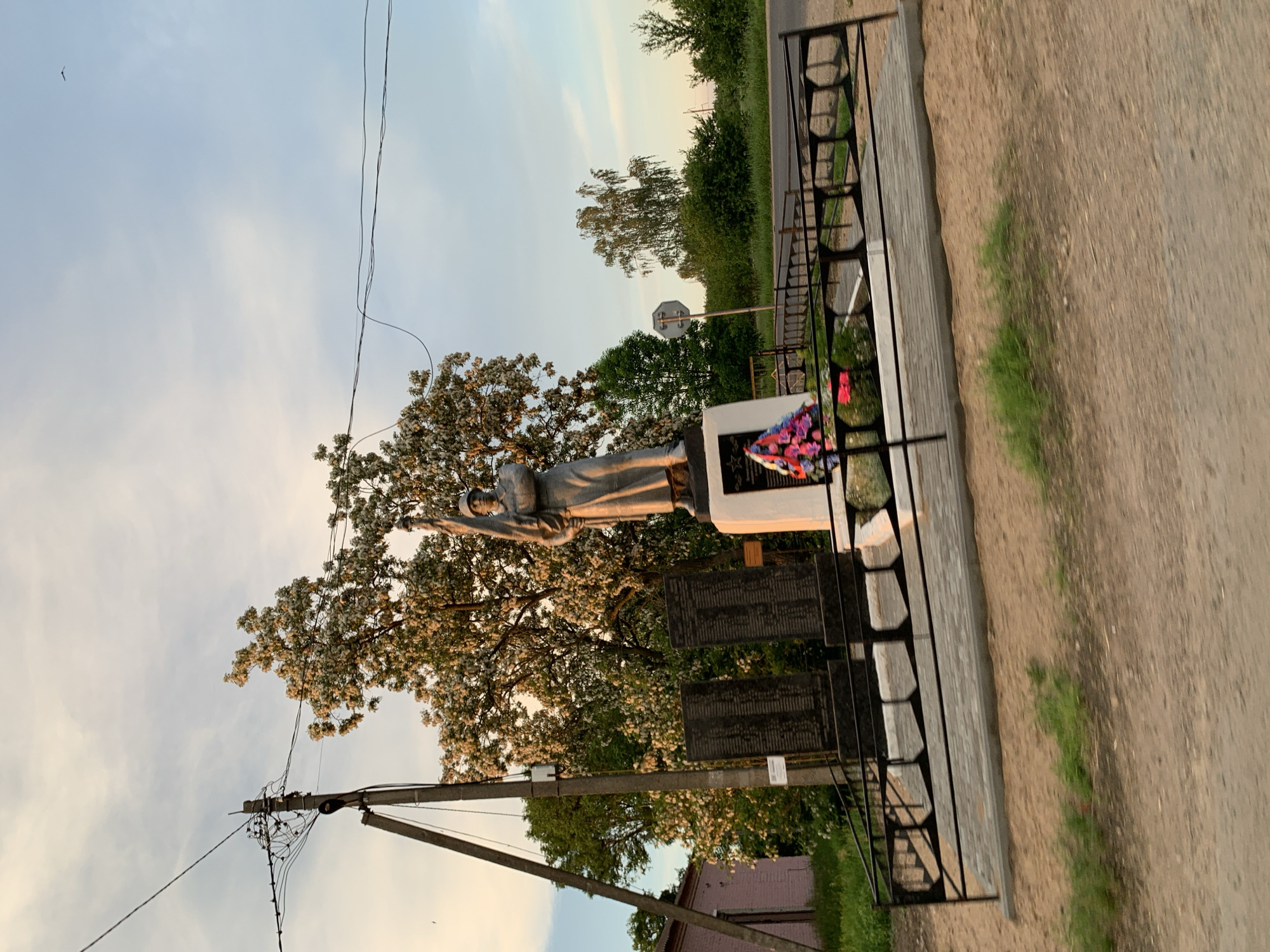 The image depicts a war memorial located near the village of Blevchitsy, within the Blevchitsy rural council of Belarus. The memorial features a statue of a soldier holding a flag, standing on a pedestal. Two dark stone tablets with inscriptions are situated in front of the pedestal, to the left. A wreath of colorful flowers is placed at the base of the pedestal. The memorial is enclosed by a black metal fence. Behind the memorial, a large tree with white blossoms is prominent. A utility pole with numerous wires is visible to the left of the memorial, and a pink building is partially visible in the far left background. The scene is set against a backdrop of trees, a road, and a sky with scattered clouds, suggesting an outdoor, rural setting during either sunrise or sunset due to the warm lighting.