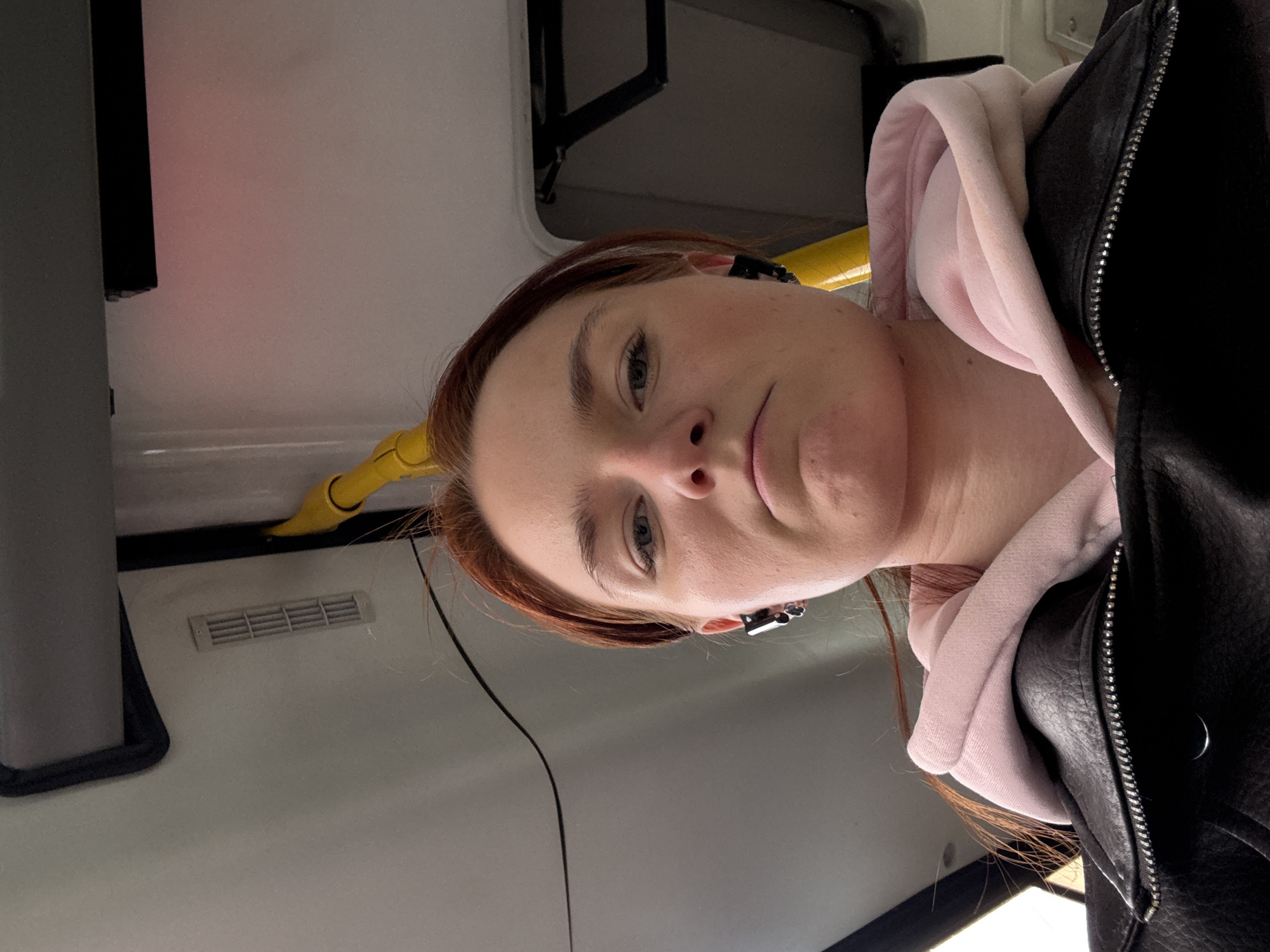 A woman with red hair and fair skin wearing a pink hoodie and black leather jacket is in what appears to be the interior of a public transportation vehicle. The vehicle features yellow handrails and gray interior panels. The location in the city of Saint Petersburg, Russia, is not discernible from the image.