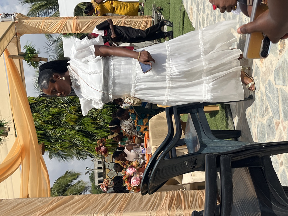 The image portrays an outdoor social gathering in Kumasi, Ghana. In the center-right foreground, a dark-skinned woman stands on a variegated stone-paved surface. She is wearing a white, floor-length, tiered, semi-sheer dress with ruffled short sleeves, a high collar, and horizontal dark gray bands on the bodice. Her dark hair is styled in a high ponytail with laid edges, and she wears pearl drop earrings. A thin necklace is visible, and she holds a dark-cased smartphone in her right hand. Gold-colored, embellished high-heeled sandals are on her feet, and a small patterned shoulder bag with a gold chain strap is visible on her left hip. She is facing slightly left, with her head turned to the left.
To the left, several dark plastic chairs are arranged near a round table covered with an orange tablecloth. Two floral centerpieces, containing pink, orange, and maroon flowers, are on the table alongside glassware and a water bottle. Multiple individuals are seated around this table, partially visible, some dressed in patterned attire.
In the mid-ground right, a man in a black long-sleeved shirt with a red and white shoulder panel and black trousers stands on artificial green turf. A woman in a yellow garment is partially visible behind him. Overhead, orange drapery fabric is stretched across a support structure, from which green leafy decorations are suspended. Lush green trees and shrubs form the background, with a stone wall visible behind the seated guests. An overcast sky is present above the foliage. In the lower right foreground, a hand holding a light-colored smartphone with visible camera lenses is partially depicted.