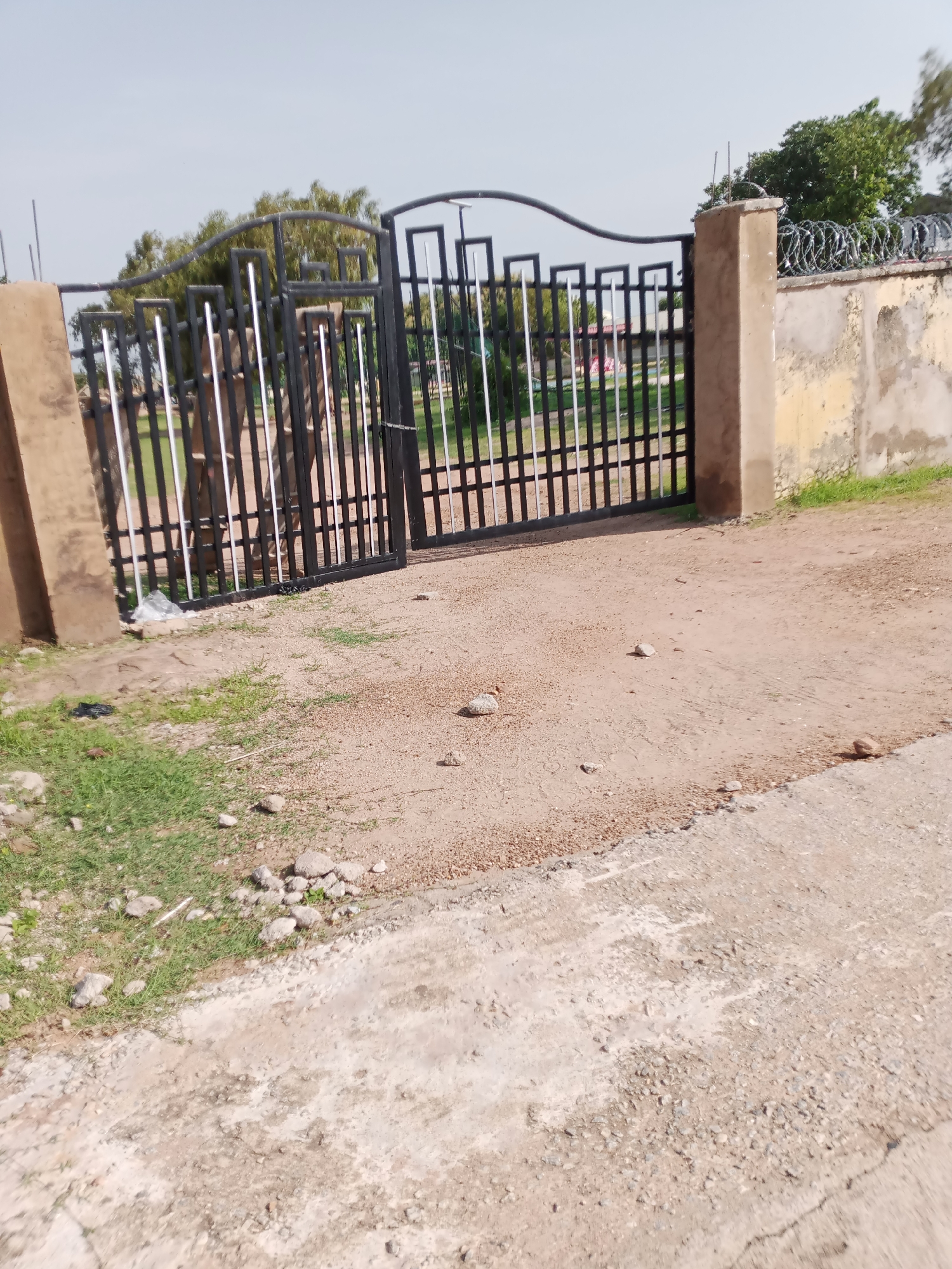 The image shows a black metal gate with a curved top, enclosing a section of what appears to be a park or recreational area. The gate is supported by a thick, light-brown concrete pillar on the left and a similar pillar on the right, which is topped with coiled barbed wire. Behind the gate, trees and some structures, including a building with a pink roof, are visible. The ground in front of the gate is a mix of dirt and gravel, with patches of green grass on the left side of the image. The area appears to be an outdoor space within a city.