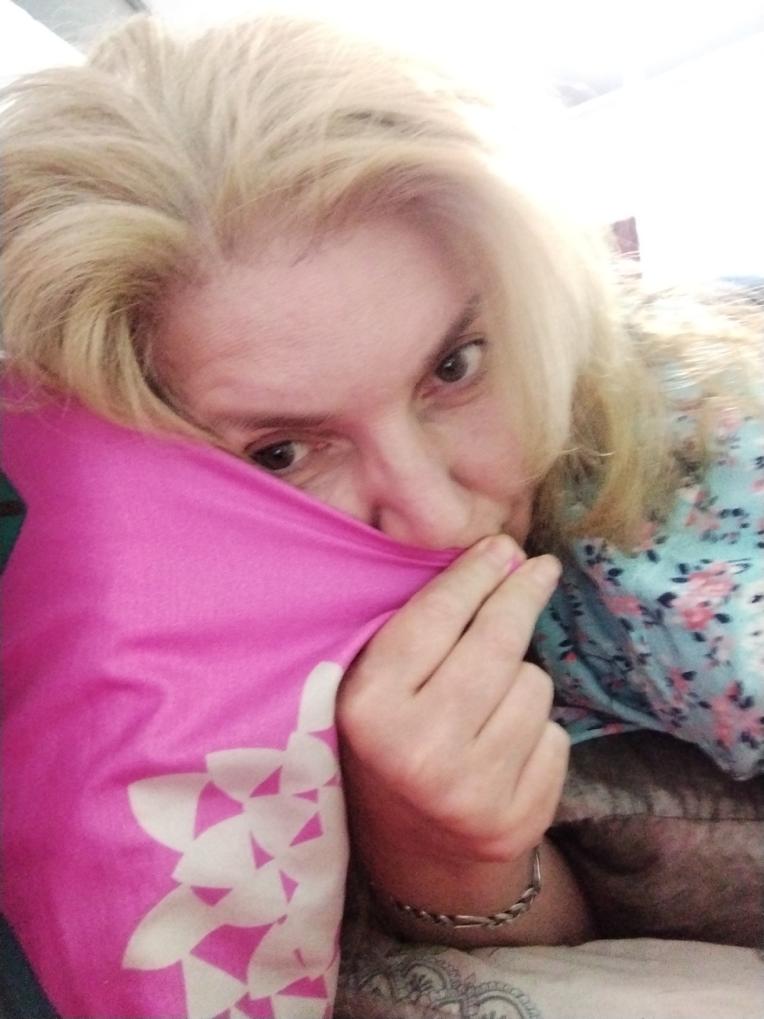 A woman with blonde hair is resting her head on a pink pillow with a white floral pattern. She is wearing a blue top with a floral print. Her right hand is resting on the pillow, and her left arm is visible on the right side of the image. She appears to be in a bedroom. There is no indication of the location being in Al Mas'oudi, United Arab Emirates.