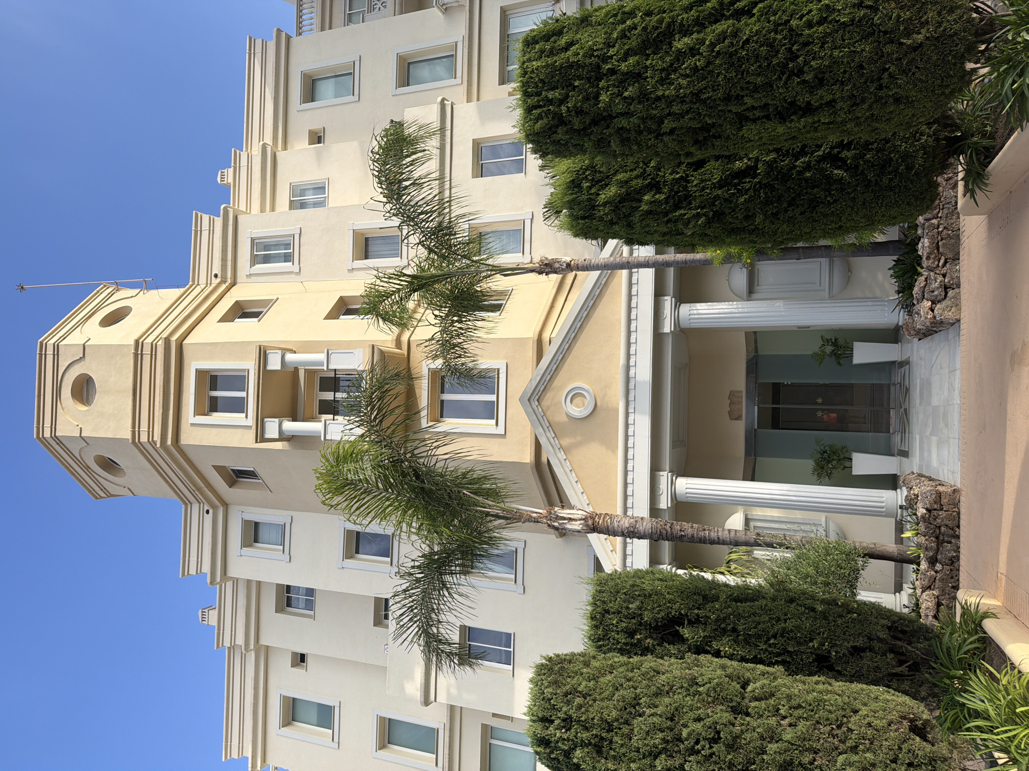 The image captures the facade of a cream-colored apartment building in Estepona, Spain, under a clear blue sky. The building features a prominent octagonal tower with circular decorative elements, topped with a spire. White-framed windows are arranged symmetrically on its multiple levels. The entrance is flanked by large white columns and leads to double glass doors. Lush green cypress trees frame the entrance, with a palm tree in the foreground. Two white planters, each with a small green plant, are positioned on either side of the entrance pathway. The ground is paved with light-colored tiles. The scene appears serene and well-maintained.