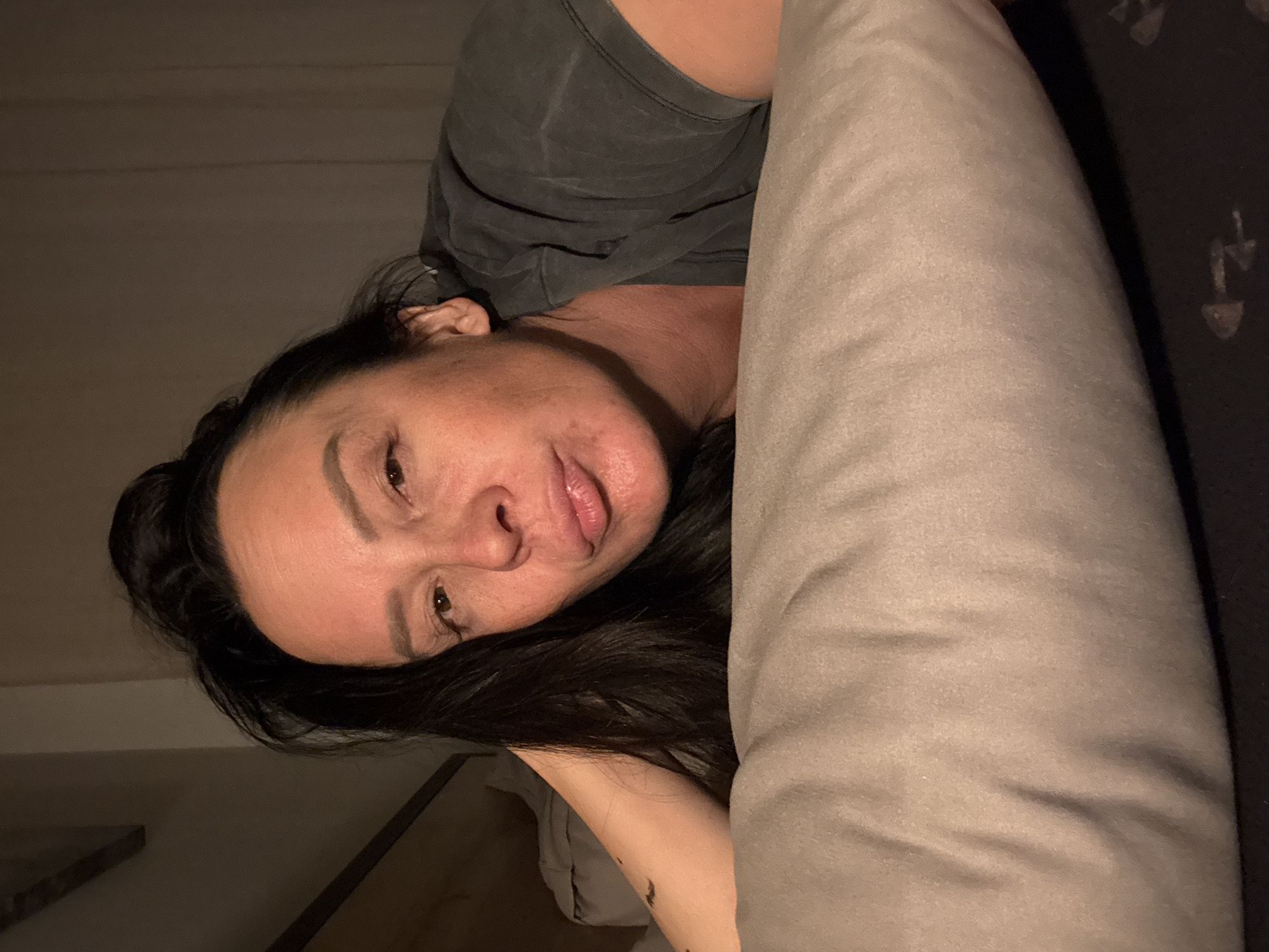 A person is lying down in what appears to be a bed or cushioned surface. The person has dark hair and is wearing a dark-colored shirt. A large pillow or cushion is positioned near the person's face. The background appears to be an interior space, possibly a bedroom, with a wall partially visible. The lighting is subdued, and the orientation of the image is rotated. There is no visible information to determine a location in the city of Chaikovsky, Russia.
