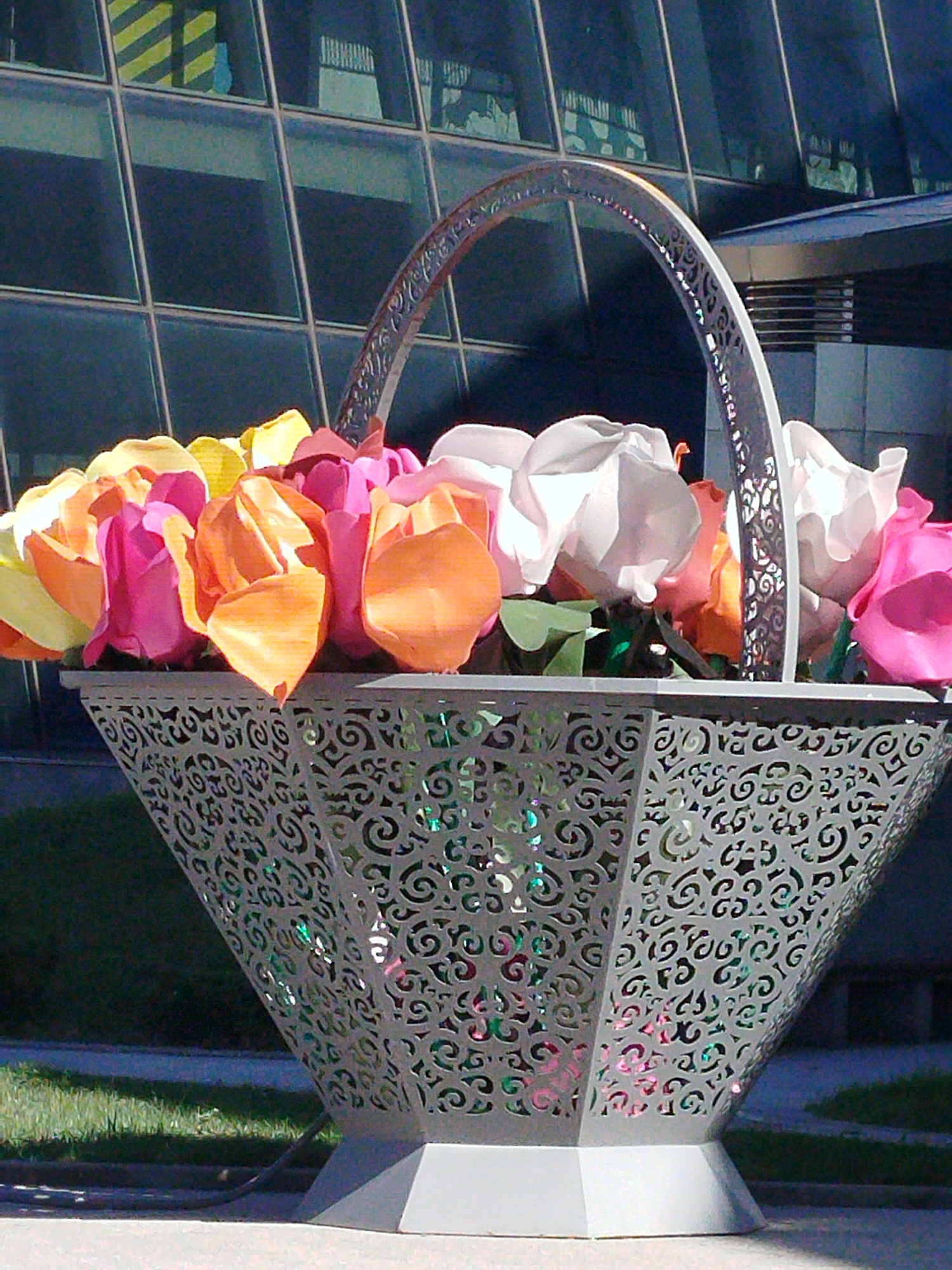 A large, decorative basket filled with colorful, artificial flowers is displayed outdoors in front of a modern glass building. The basket itself is made of intricate, silver metalwork with a lattice pattern and a curved handle. Inside the basket, the flowers are predominantly pink, orange, and yellow, with some white and light pink ones mixed in. The building in the background features a grid of large windows, reflecting the sky and surrounding environment. The scene appears to be a public space, possibly a plaza or entrance to a building, in the city of Astana, Kazakhstan. The ground in front of the basket is paved, and grass is visible to the left. The lighting suggests it is daytime, with sunlight casting shadows.