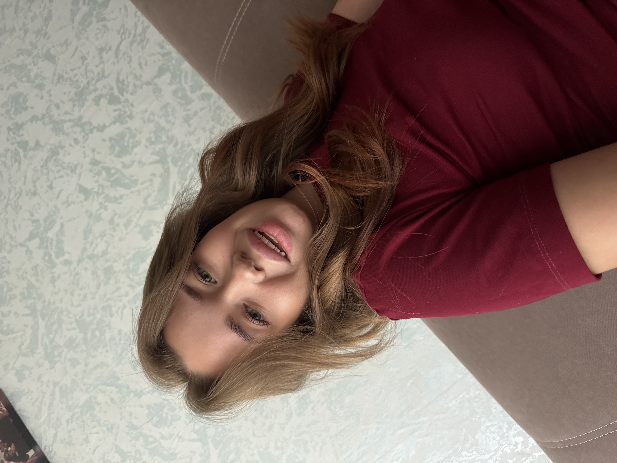 A young woman with long, wavy brown hair and green eyes smiles at the camera. She is wearing a dark red t-shirt and is lying on a brown sofa. The background appears to be a wall with a light blue and white patterned wallpaper. The image is a close-up shot, focusing on the woman's face and upper body. There is no specific indication of the location within Chaikovsky, Russia.