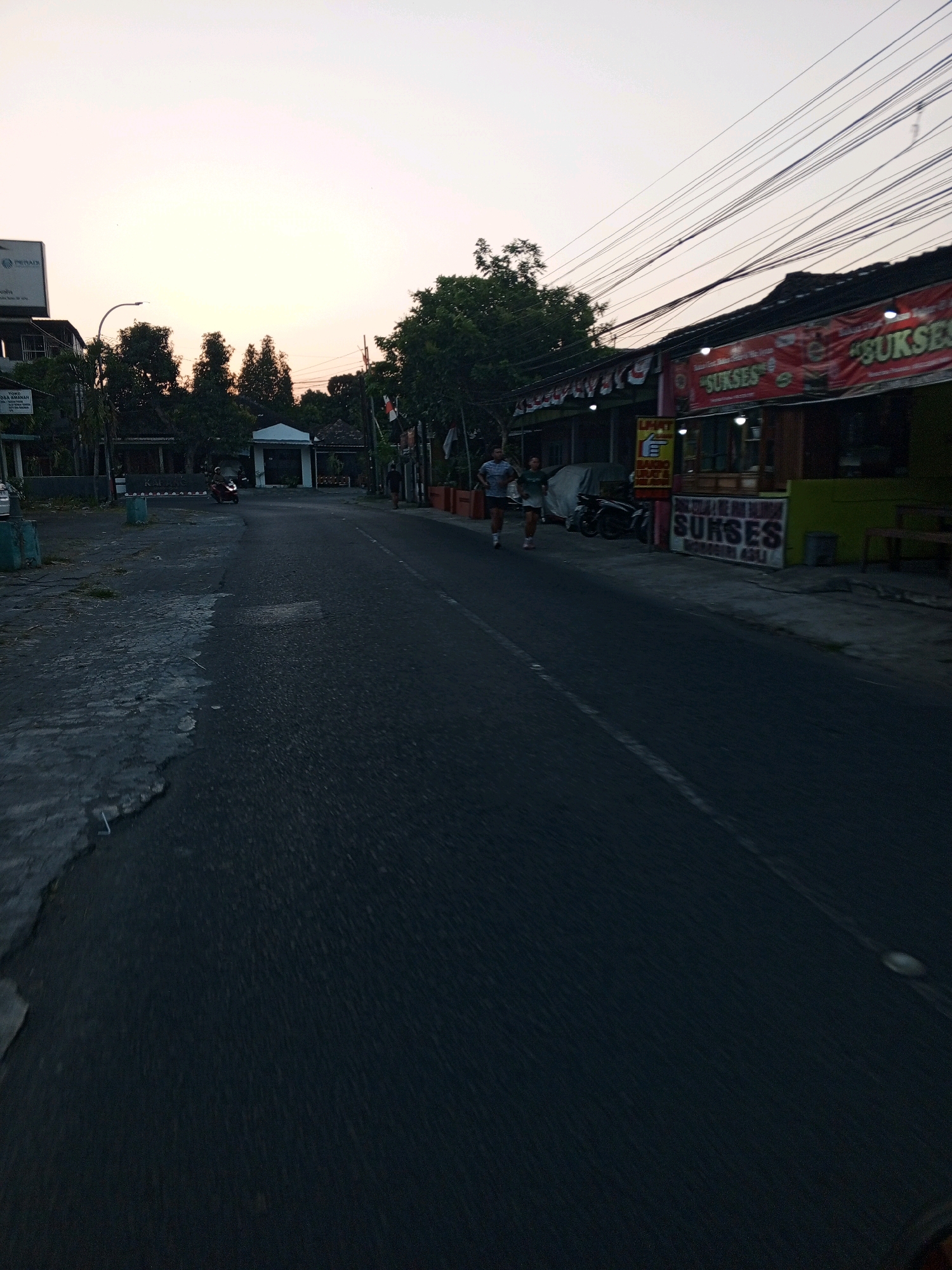 The image depicts a street scene in Bantul, Indonesia, during what appears to be early morning or late afternoon, evidenced by the soft, warm lighting and pale sky. In the foreground, a paved road with a white line marking the lane occupies a significant portion of the frame. The left side of the road features a damaged concrete or asphalt curb, with debris and patches of growth.
Further down the road, two individuals are jogging, one wearing a blue shirt and white shorts, the other a white shirt and green shorts. To the right of the road, a row of buildings lines the street. One building prominently displays a red banner with the word "SUKSES" repeated multiple times in white text, suggesting a commercial establishment. There are other signs with Indonesian text, and several motorcycles are parked in front of the buildings. Trees are visible behind the buildings, and utility wires crisscross the sky above. In the distance, a white structure resembling a small pavilion or gate is situated near a cluster of trees, and a motorcycle rider is approaching it. The overall atmosphere is tranquil, suggesting a typical day in a semi-urban Indonesian environment.