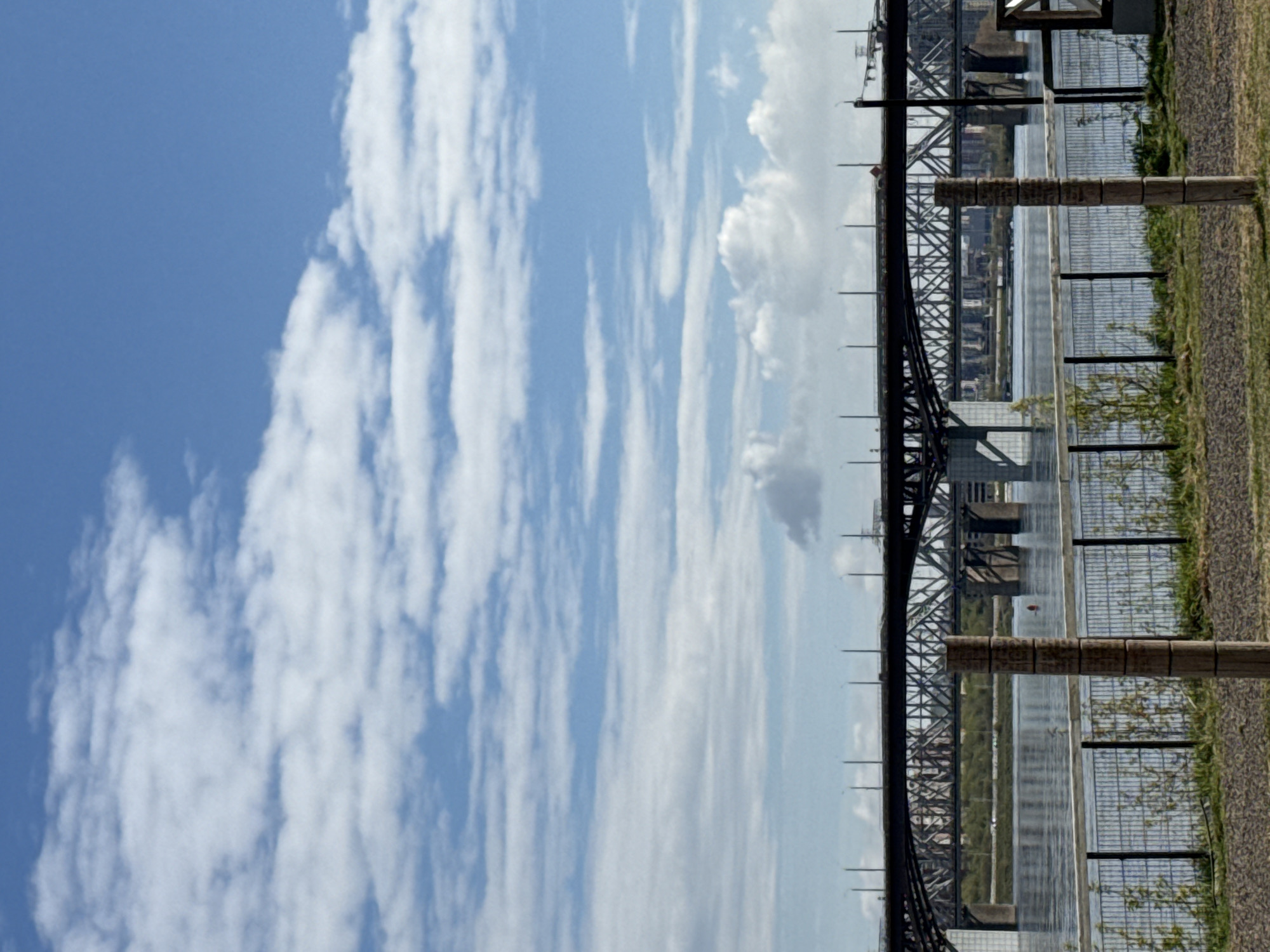 The image shows a view of the Yenisey River in Krasnoyarsk, Russia, with a steel truss bridge spanning across it. To the right of the bridge is a sidewalk with a barrier fence, and beyond that, a strip of green vegetation. The river reflects the sky, which is predominantly blue with elongated white clouds streaking across it. The bridge's steel framework is visible in detail, suggesting an industrial design. There are no people visible in the scene.