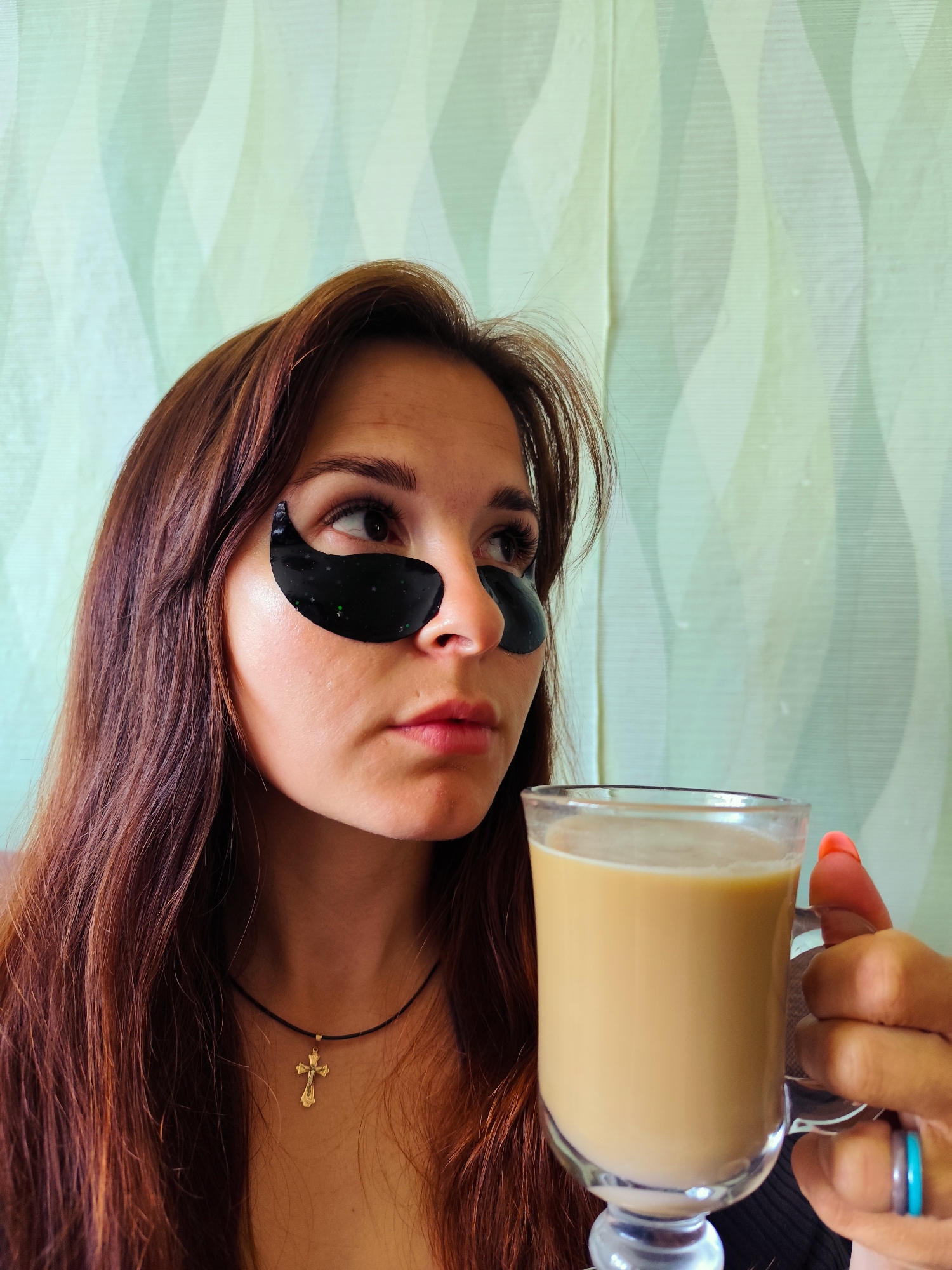 A close-up, medium shot shows a young woman with long, dark brown hair and black under-eye patches, holding a clear glass mug of coffee with milk. She is looking to her right with a neutral expression. The woman wears a thin black necklace with a gold cross pendant. Her left hand, with bright pink fingernails, is visible holding the mug, and she wears two thin rings on her index finger: one blue and one clear. The background is a light green, wavy patterned curtain. The image does not provide any visual cues that would allow for the identification of the location in the city of Fokino, Russia.