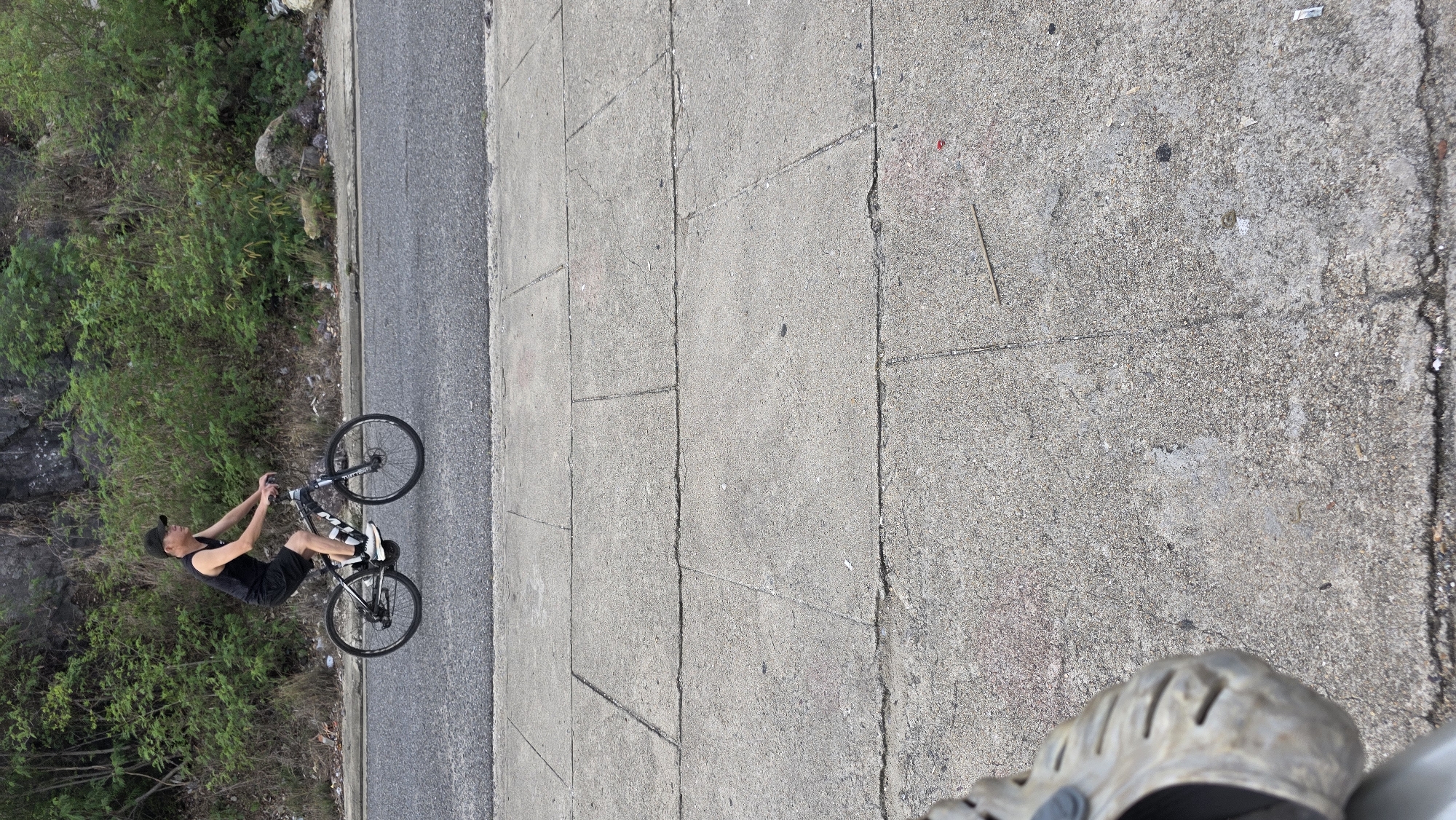 This outdoor image, captured in Bac Nha Trang Ward, Vietnam, presents a man on a bicycle engaged in a wheelie. The photograph is taken from a slightly elevated perspective, looking down, with the photographer's foot in a grey Croc-style sandal visible in the bottom right corner.
The central subject is a man wearing a black tank top, black shorts, a black baseball cap, and white sneakers. He is balanced on the rear wheel of a black bicycle, with the front wheel lifted high in the air, demonstrating a wheelie. The brand name "KONA" is visible on the bicycle's frame.
The man is riding on an asphalt road. Adjacent to the road is a concrete path or sidewalk, which shows cracks and scattered small debris like a red item, a wooden stick, and tiny pieces of paper. To the left of the road and path, there is a verdant, densely vegetated area, likely a hillside, covered in green bushes and trees, with some exposed rocks among the foliage.
The scene is well-lit, indicating it is daytime with bright, clear weather. The setting combines a paved pathway and road with a natural, overgrown hillside, suggesting a roadside area perhaps on the outskirts or through a natural park in the region.