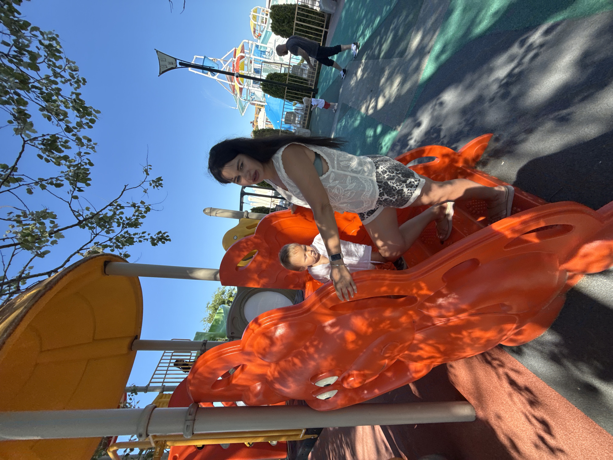 In Sochi, Russia, on a sunny day, a woman and a toddler are on a playground. The woman, with long dark hair, wears a white lacy top and patterned shorts. She is positioned on an orange dragon-shaped slide. The toddler, wearing a white shirt, is also on the slide, looking down. In the background, other people and water park attractions are visible, including slides and a tall structure with spraying water, suggesting the playground is part of a water park or amusement area. The ground is a mix of textured surfaces in grey, orange, and green. Trees with green leaves are overhead, casting shadows on the ground.