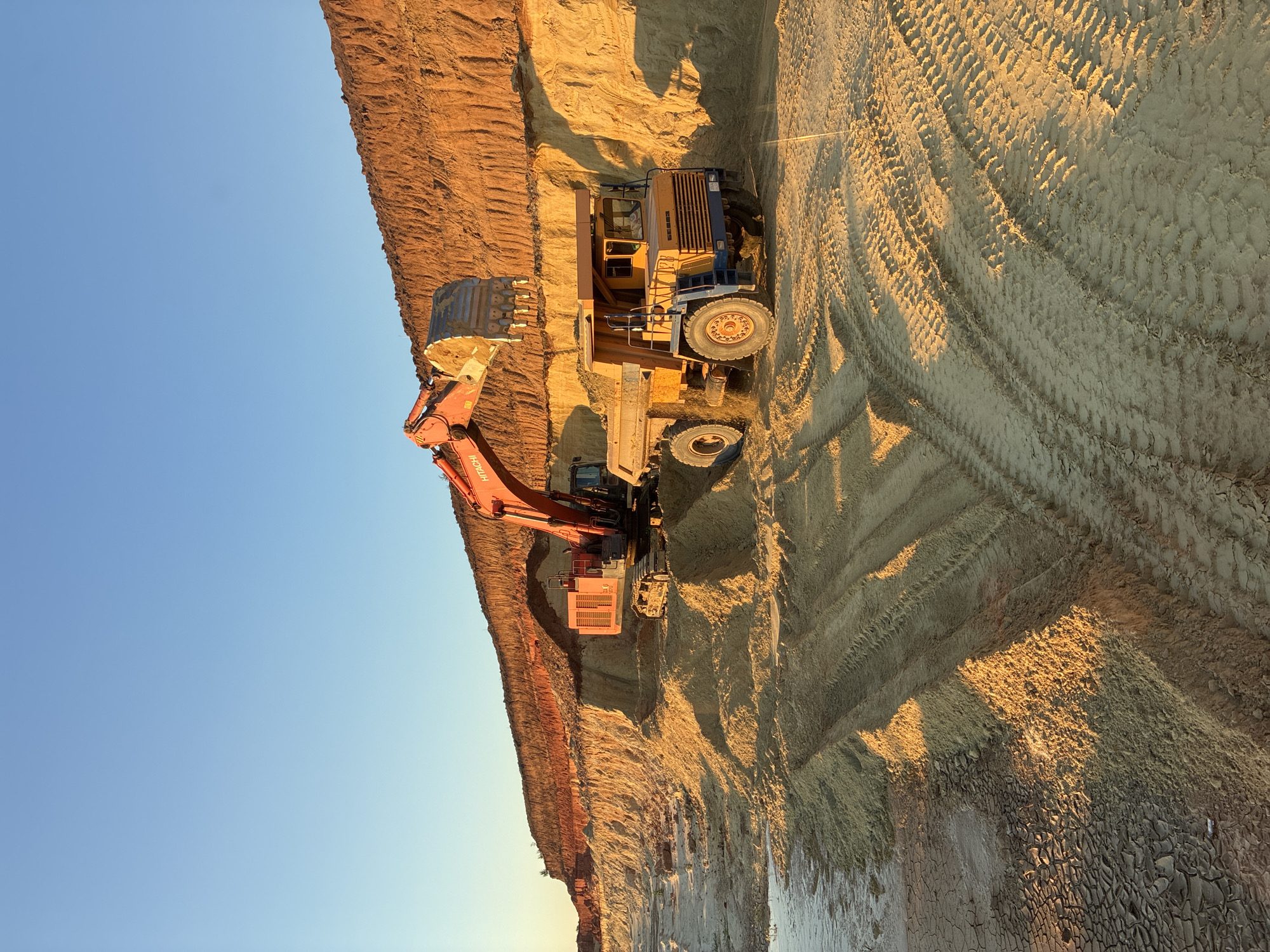 In an outdoor mining or construction setting, likely in Tatsinskaya, Russia, a large orange Hitachi excavator is actively digging and loading earth into a yellow and blue dump truck. The excavator's bucket is full, and it's in the process of dumping its load. The dump truck is positioned to receive the material, with its bed open. The scene is set against a clear blue sky, suggesting fair weather. The warm, golden light illuminates the excavated earth, casting long shadows and indicating that it is either early morning or late afternoon, with the sun low on the horizon. The ground is heavily textured with tire tracks from heavy machinery, signifying ongoing activity. The excavated earth shows distinct layers of different soil colors, ranging from reddish-brown at the top to lighter sandy tones lower down. The overall impression is one of active, large-scale earthmoving operations. Visible text on the excavator reads "HITACHI".