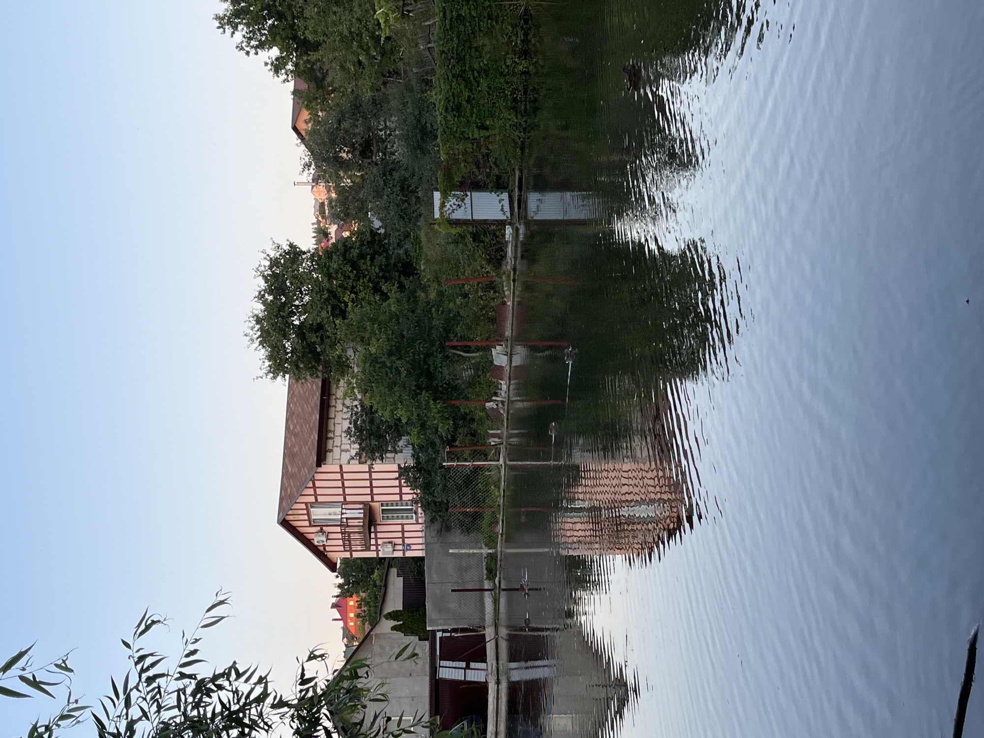 The image depicts a serene waterscape with a house and surrounding foliage reflected in the water. The house is a two-story structure with a distinctive pink and white facade, featuring a balcony on the upper floor and a reddish-brown tiled roof. It appears to be situated in a suburban or rural setting, given the presence of trees and other residential buildings in the background. Several ducks are visible in the water, swimming peacefully. The water itself is calm, with gentle ripples distorting the reflections of the house and the sky. The overall atmosphere is tranquil and evokes a sense of quietude. The image could be from the city of Saratov, Russia, as indicated by the prompt.