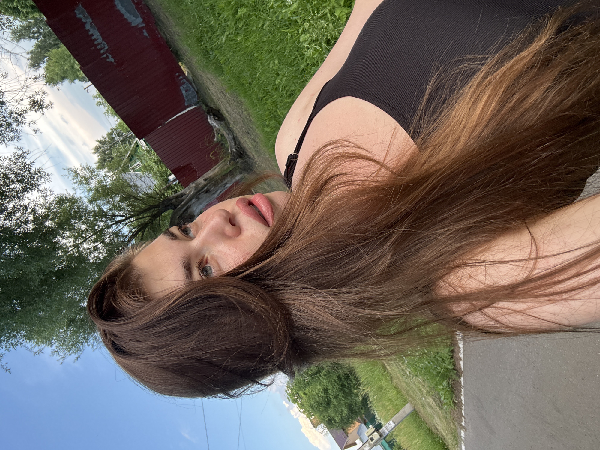 The image shows a young woman with long, brown hair, wearing a black tank top. She is outdoors in a grassy area with a red fence and trees in the background. The sky is visible with some clouds. The woman is looking off to the side, and her face is partially obscured by her hair. The location is identified as Мещёры, Russia.