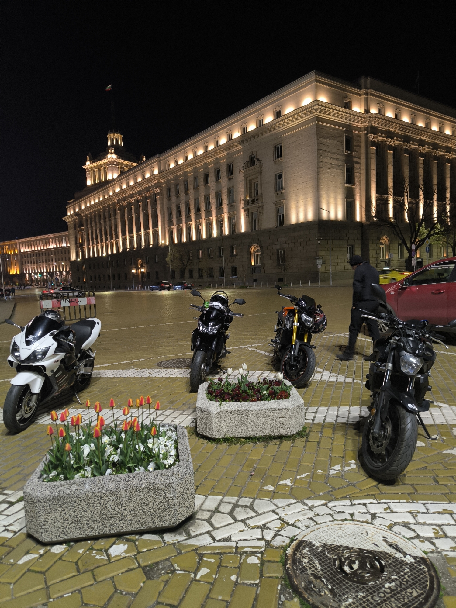 The image presents a nighttime street scene in Sofia, Bulgaria, dominated by a large, ornate building and several parked motorcycles. The time is clearly night, with the sky being dark, and the building extensively illuminated by warm spotlights that highlight its classical architecture, including numerous columns and detailed cornices. A Bulgarian flag, with its white, green, and red stripes, flies from a flagpole atop a central tower of the main building. Another grand building is visible in the background, also lit.
In the foreground and mid-ground, at least four motorcycles are parked on a plaza paved with distinctive yellow-orange and white geometric stone blocks. A white Honda motorcycle is visible on the left, with the brand name "HONDA" clearly legible. Other motorcycles are black or black with orange accents. A person, dressed in dark clothing, is seen walking among the parked motorcycles on the right side of the frame.
Several concrete planters, filled with red and yellow tulips and white flowers, are interspersed among the motorcycles. In the lower right corner, a manhole cover, partially painted white, is visible on the paved ground. Cyrillic text on the manhole cover reads "СОФИЯ" (Sofia) and "ДВУК 1680-70". The overall scene captures a quiet urban evening against a backdrop of impressive, historically significant architecture.