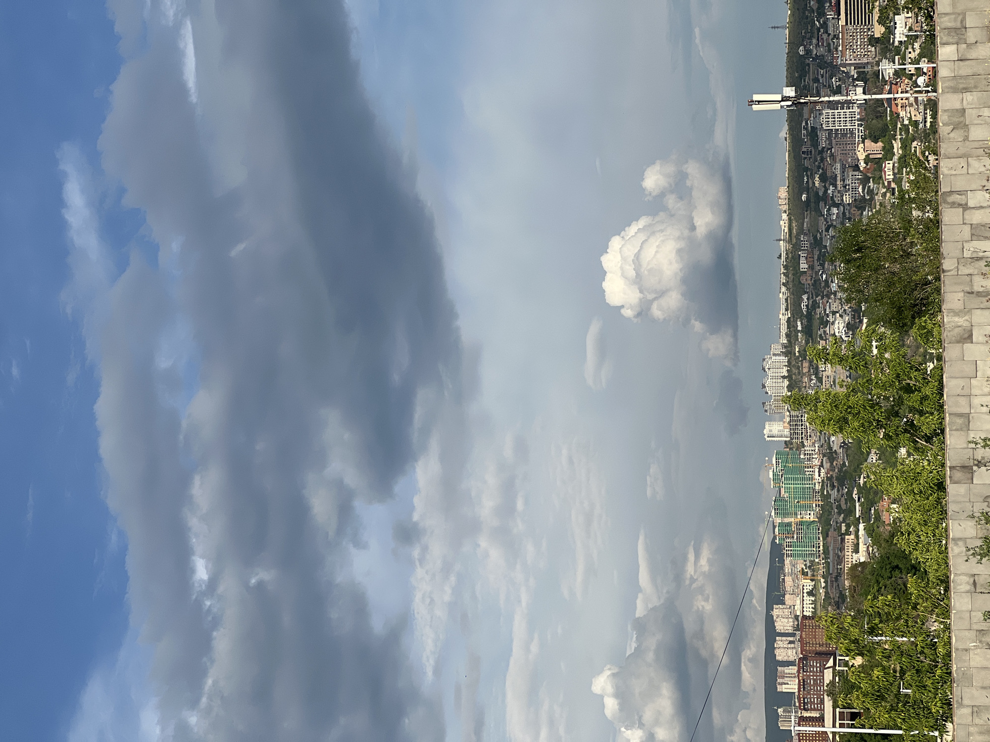 The image presents a panoramic elevated view of Yerevan, Armenia, under a dynamic sky. The upper two-thirds of the frame are dominated by a blend of dense, dark gray stratocumulus clouds and lighter, fluffier cumulus formations, with patches of clear blue sky visible, particularly in the upper-left and upper-right portions.
In the foreground, a low, segmented wall constructed from light gray and beige rectangular blocks runs horizontally across the bottom. Directly behind this wall, a line of vibrant green deciduous trees and bushes with dense foliage is visible, partially obscuring the immediate cityscape.
The mid-ground reveals a sprawling urban landscape characterized by a dense concentration of buildings spread across undulating terrain. Numerous residential structures of varying architectural styles and ages are present, ranging from individual houses with light-colored roofs to multi-story apartment blocks. Several clusters of modern high-rise buildings, some appearing to be under construction and covered with distinctive green protective netting, are prominent. A slender, dark power line traverses diagonally from the upper-left corner of the cityscape towards the center-left. On the far right, a tall, white cellular communication tower equipped with multiple antenna panels stands prominently above the residential areas.
The background features distant hills or low mountains that form the horizon line, partially veiled by atmospheric haze and the expansive cloud cover. No people or specific actions are discernible within the frame. The lighting suggests daylight conditions, with a mix of direct and diffused light due to the partial cloud cover.
Based on the panoramic urban view, the distinctive topography, and the prevalence of both older and newer residential developments, the image is captured from an elevated position within Yerevan. Specifically, the vantage point is consistent with a view from **Victory Park (Haghtanak Park)**, looking generally south-southwest across the city, encompassing areas of both central Yerevan and newer developments on the surrounding hills.