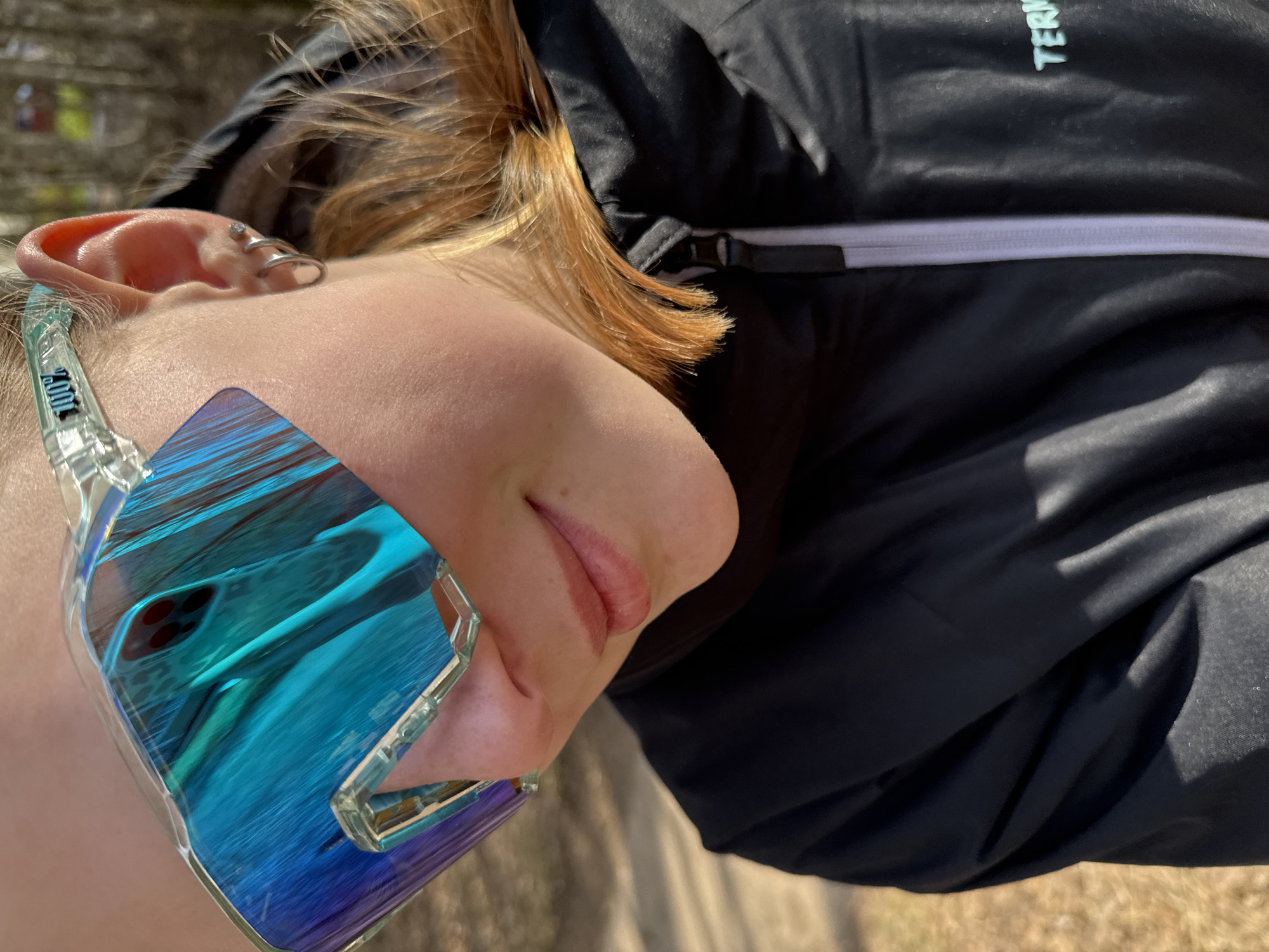 This close-up, tilted shot captures the upper body of a person outdoors in bright sunlight. The individual has light brown or auburn hair that falls across their shoulder and neck. They are wearing a dark, possibly black, jacket with a visible zipper and a strap with a buckle. Partial text, "TERM", is visible on the jacket, likely part of a brand name.
The person's right ear is adorned with several silver-colored piercings, including two helix rings and a small stud. Mirrored, blue-tinted sunglasses with clear frames are worn, reflecting what appears to be the back of a smartphone with multiple camera lenses, set against a blue, textured background. The brand "100%" is visible in white text, inverted, on the clear arm of the sunglasses. The visible part of the person's face, including their mouth and chin, shows a relaxed, subtle smile. The strong lighting suggests it is daytime with sunny weather. The background is softly blurred but indicates an outdoor environment with natural ground. This scene is set in Chaikovsky, Russia.