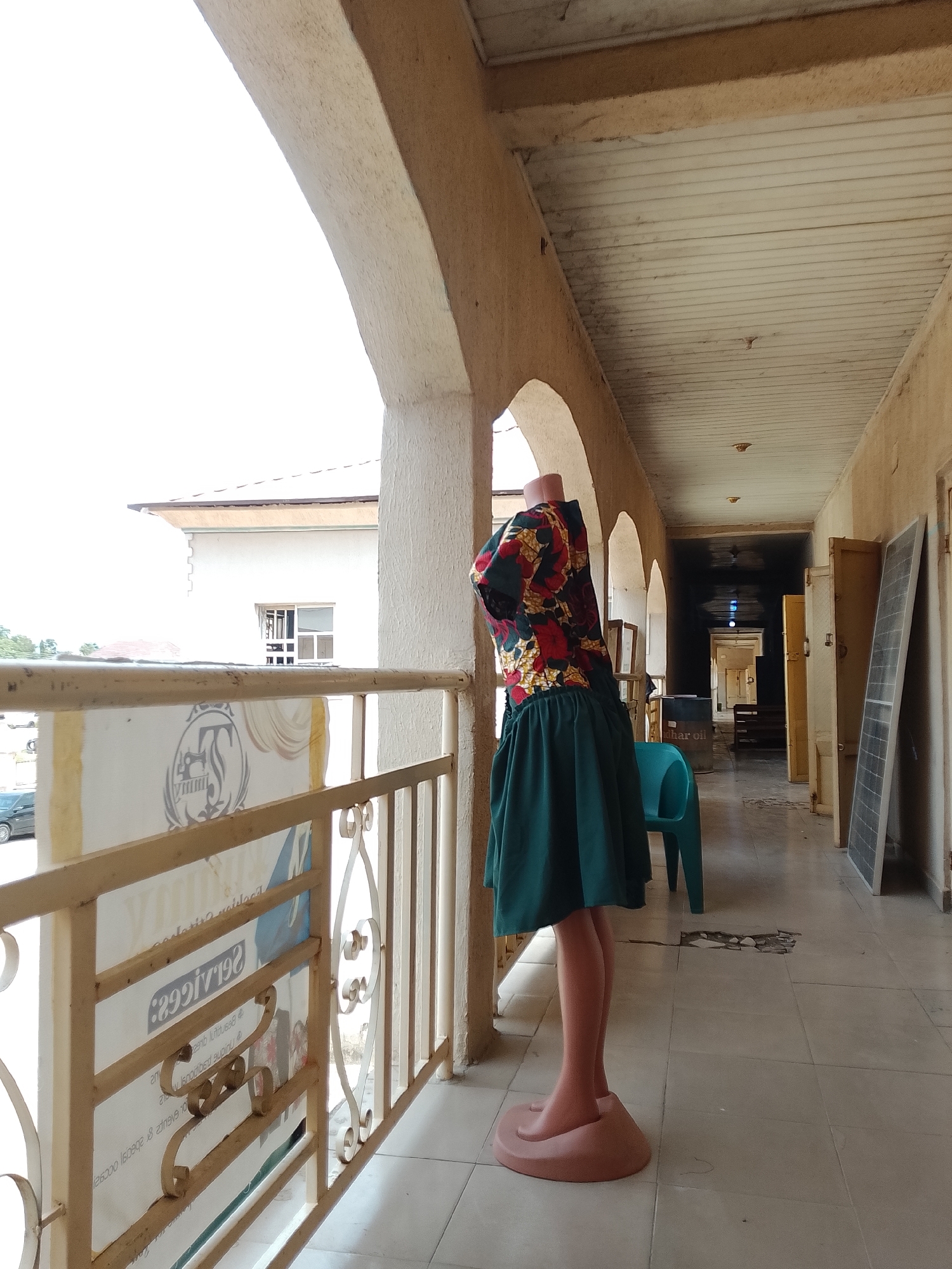 The image shows a mannequin dressed in a colorful floral print top and a green skirt, standing on a balcony or veranda. The mannequin is positioned to the left, facing away from the camera and looking out over a railing. Behind the mannequin, a long, open-air corridor stretches into the distance, with several doors on the right side. The corridor is lined with what appear to be storage cabinets or doors, and there are solar panels leaning against one wall. A turquoise plastic chair is visible near the mannequin. The floor is tiled, and there are some cracks and debris on the floor in the distance. The ceiling of the corridor is made of white wooden planks.
In the background, through the open archway to the left, a white building with windows is visible, suggesting an urban or semi-urban environment. The sky appears bright, indicating daylight, possibly late morning or early afternoon. The overall impression is one of a commercial or residential building with an outdoor walkway.
The sign on the railing in front of the mannequin has text that is partially visible and reversed, reading "299\/192 Services." The colors and style of the clothing on the mannequin suggest it might be a display in a shop or market. There is no clear indication of the specific time of day or weather conditions beyond the daylight. The setting appears to be in Jalingo, Nigeria.