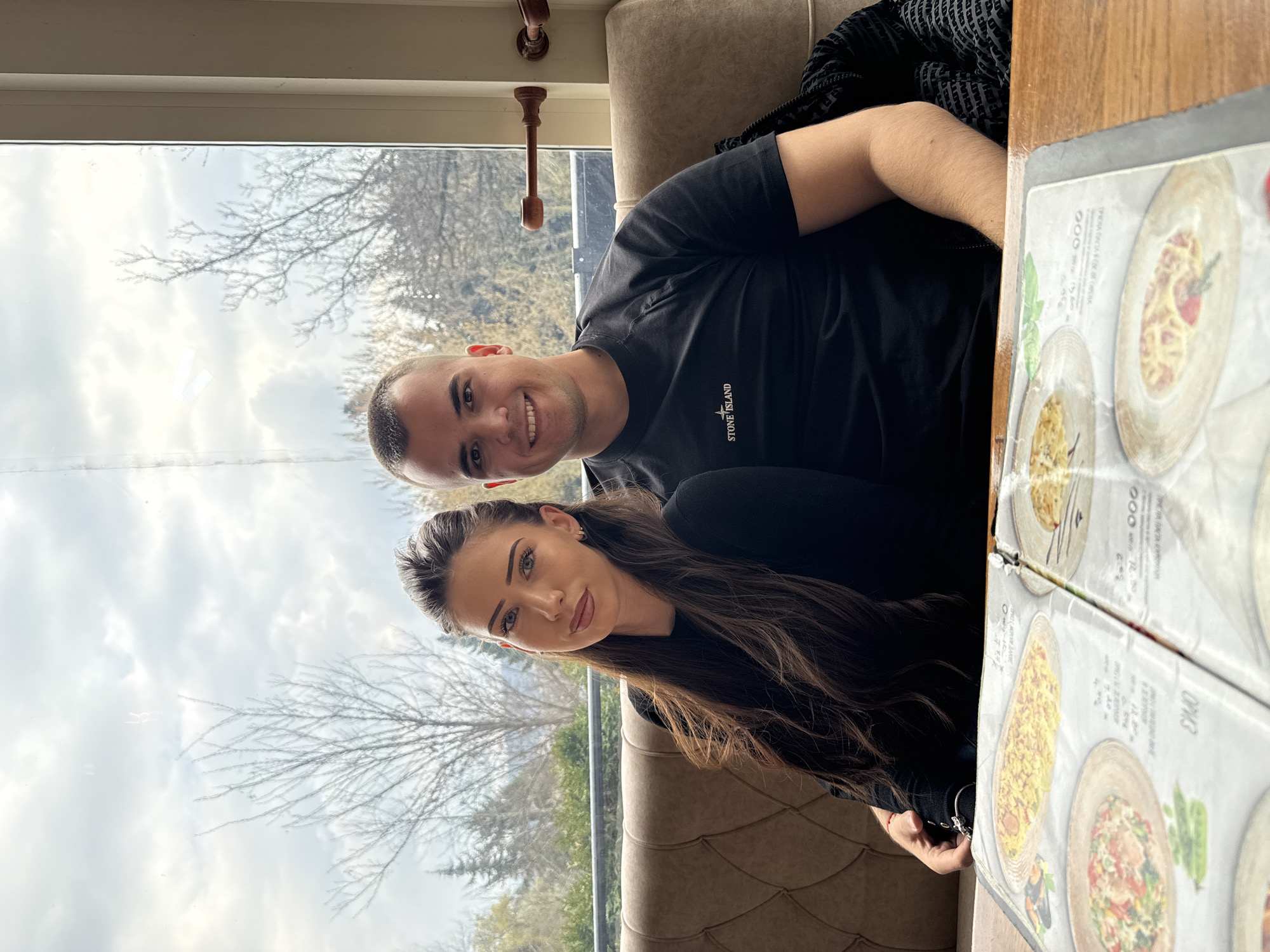 Here is a description of the image.
The image shows a couple sitting indoors near a window. The man has short hair and is wearing a black t-shirt with the words "STONE ISLAND" on it. He is smiling at the camera. The woman has long brown hair and is wearing a black top. She is also looking at the camera with a slight smile. A menu with images of food is visible on the table. Outside the window, bare trees are visible against a cloudy sky.