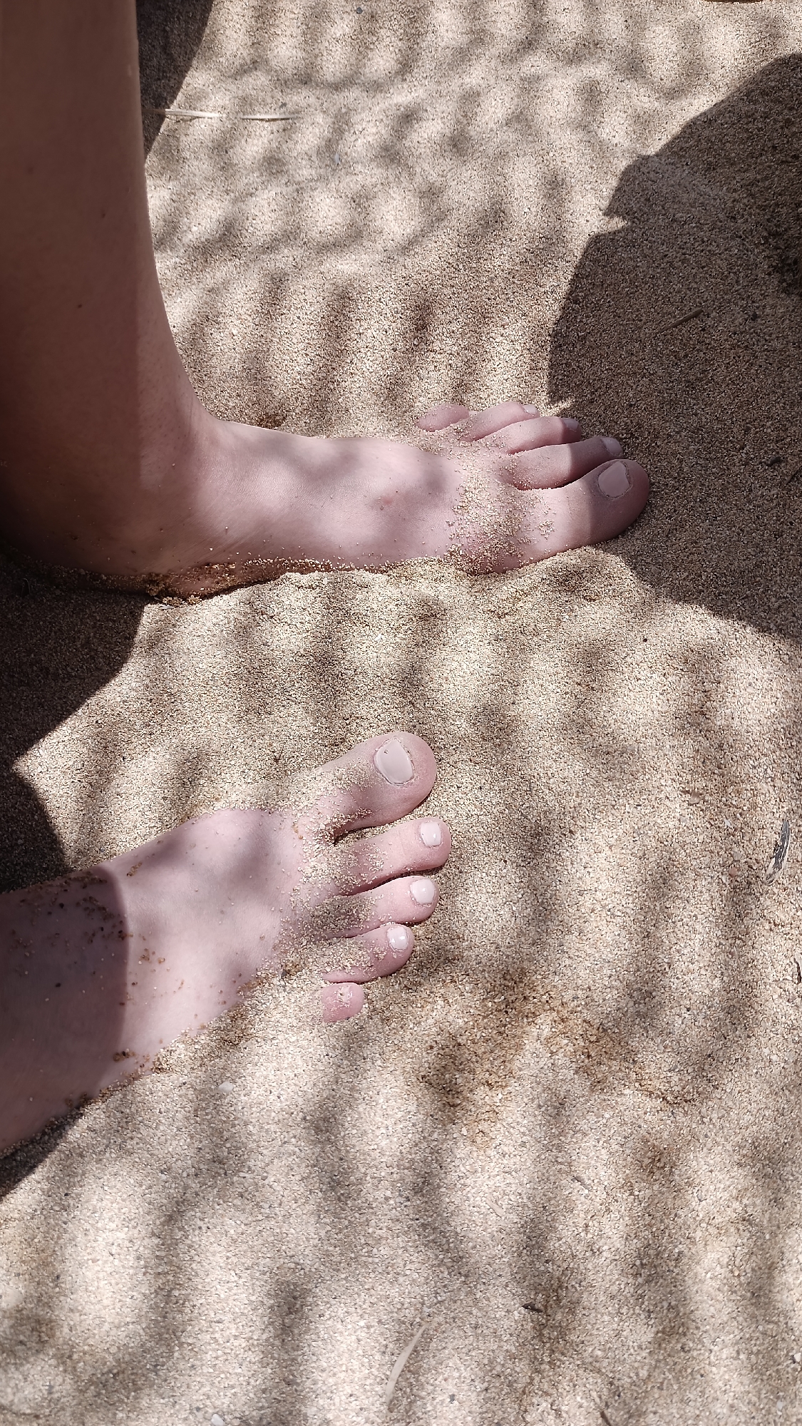 The image shows a pair of feet partially submerged in the sand on a beach. The feet are fair-skinned, and some grains of sand are adhering to them. The sand is light brown in color, and it is illuminated by dappled sunlight, creating a striped shadow pattern across the scene. The upper part of the image shows the leg of the person whose feet are visible. The location is the beach in Nea Peramos, Greece.