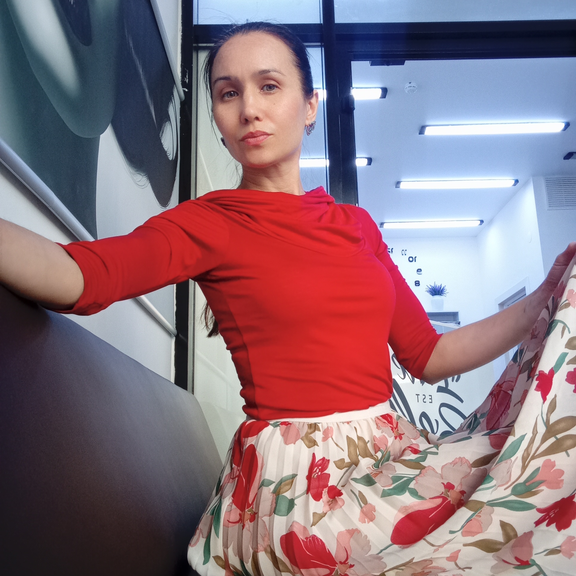A woman sits on a dark brown bench in what appears to be a brightly lit interior space in Izhevsk, Russia. She is wearing a red, long-sleeved top and a floral-patterned, pleated skirt. Her dark hair is pulled back, and she has earrings. The background features a wall with a black and white design and a window or door with multiple horizontal light fixtures visible inside. The overall lighting is bright and seems to be coming from the interior light fixtures. The setting gives the impression of a modern or recently renovated commercial space.