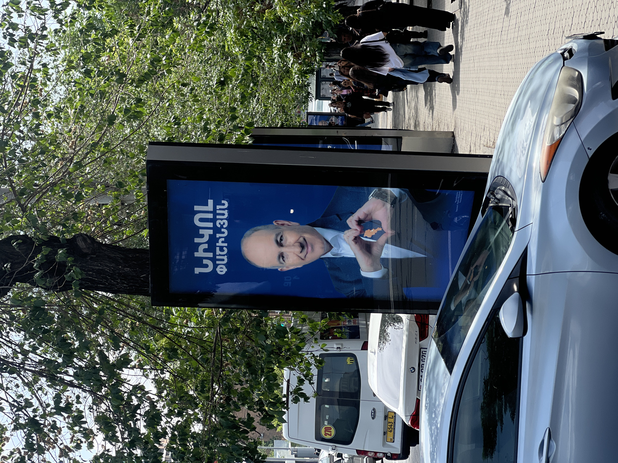 The image shows a street scene in Yerevan, Armenia. A large advertising poster is prominently displayed, featuring a man (likely a politician) against a blue background. The poster includes text in Armenian. A group of people is walking along the sidewalk in the background. Several vehicles are parked along the street, including a white van, a white BMW sedan, and a silver car partially in the foreground. Trees with green foliage are visible on the left side of the image. The scene appears to be taken during the daytime under natural lighting.