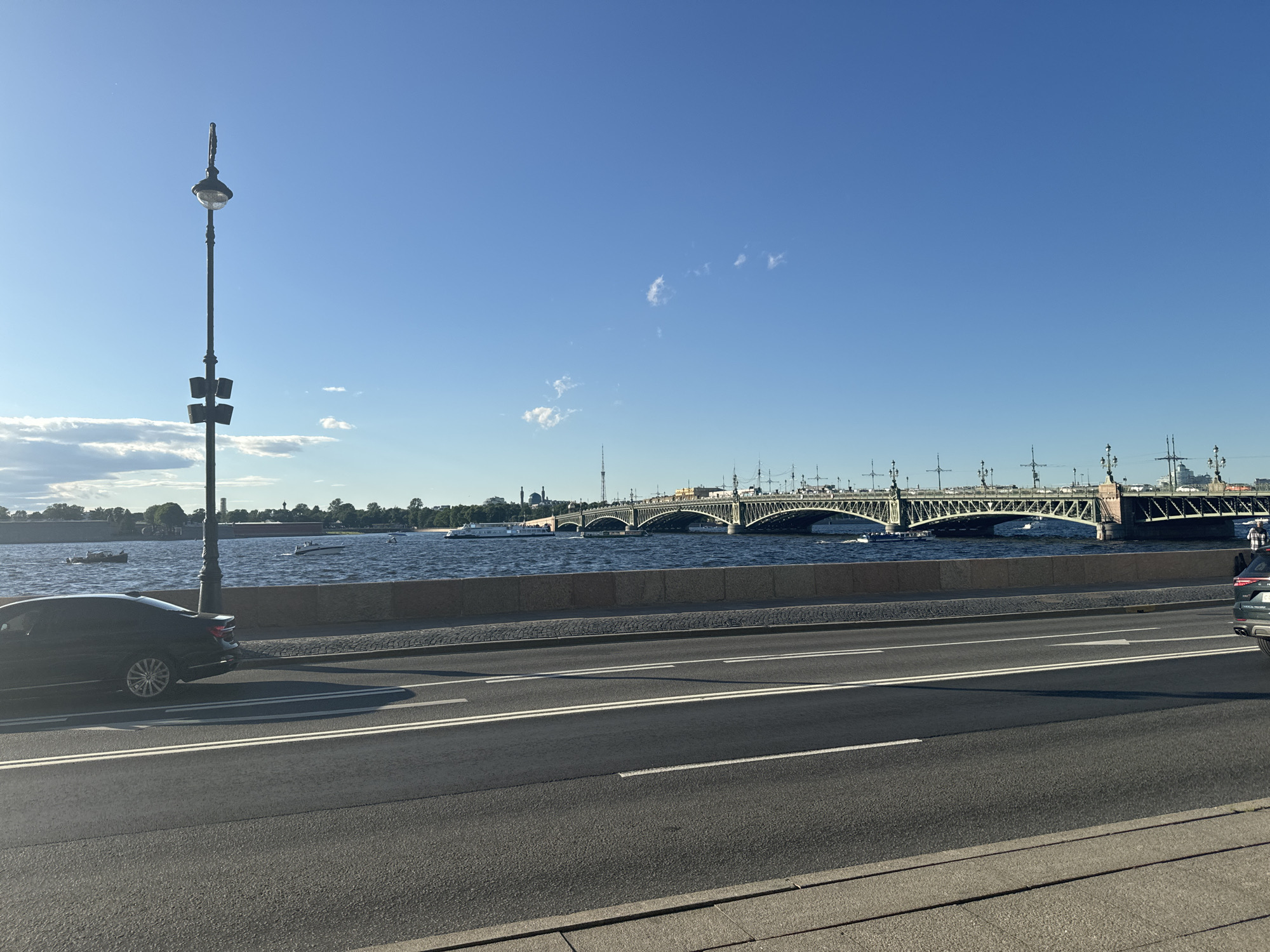 The image depicts a wide, paved road in Saint Petersburg, Russia, with a black car parked on the side and another car partially visible driving on the road. In the background, a large, green bridge spans across a wide river, with boats and ships visible on the water. The sky is a clear blue, with a few white clouds scattered across it. A tall lamppost stands on the left side of the image, overlooking the road and the river. The scene appears to be set in the afternoon, with the sun casting shadows across the road. The architecture of the bridge and the lamppost suggests a historic and grand urban setting.
