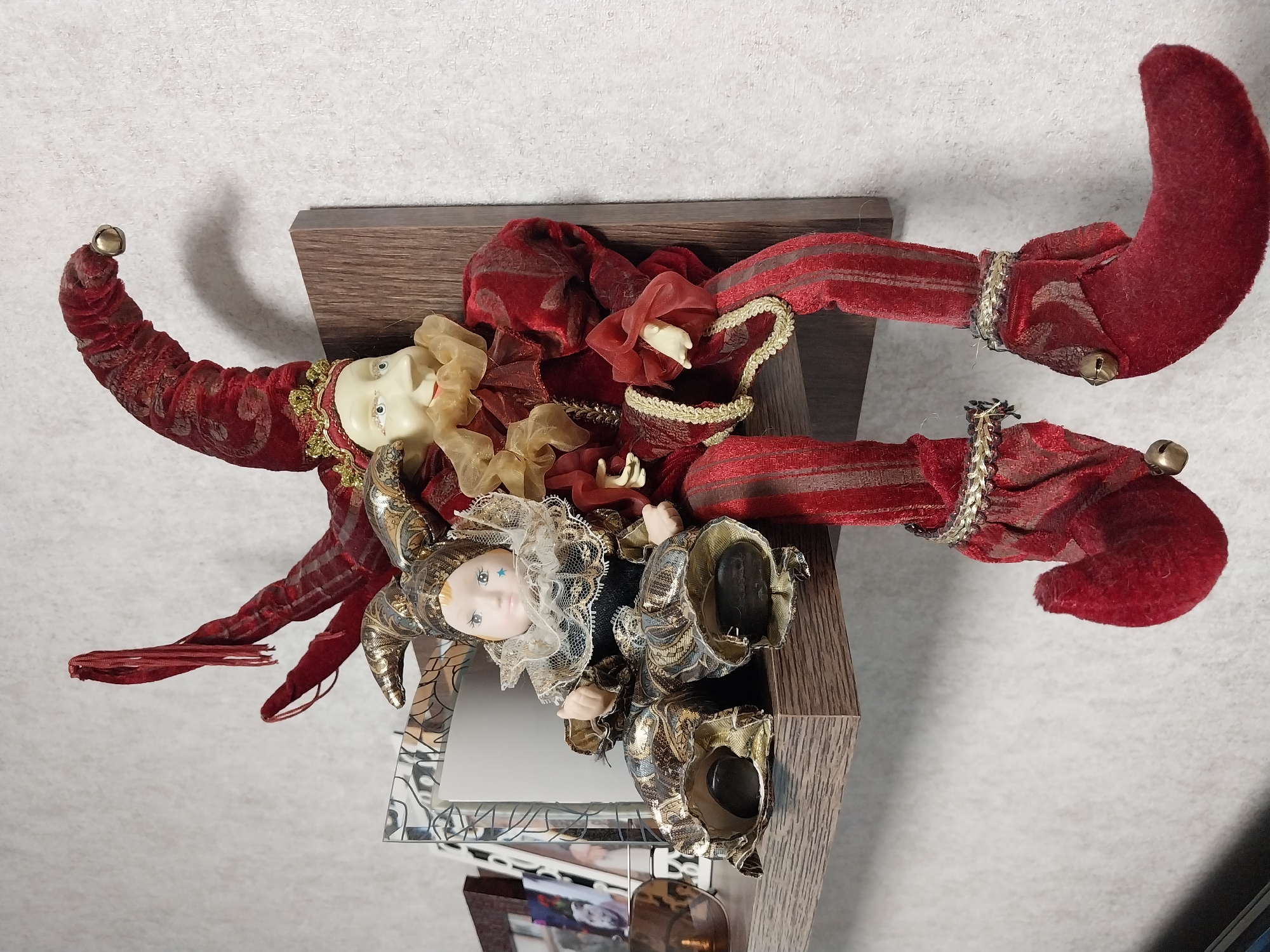 Two jester dolls are sitting on a shelf mounted on a white textured wall. The doll on the left is smaller and appears to be a baby doll dressed in a gold and black jester costume with a frilly collar. The doll on the right is larger, with a cream-colored face, red jester hat, and a red and gold costume. Both dolls have their legs dangling over the edge of the shelf. There are some picture frames and a brown object on the shelf behind the dolls. This scene is located in the city of Dnipro, Ukraine.