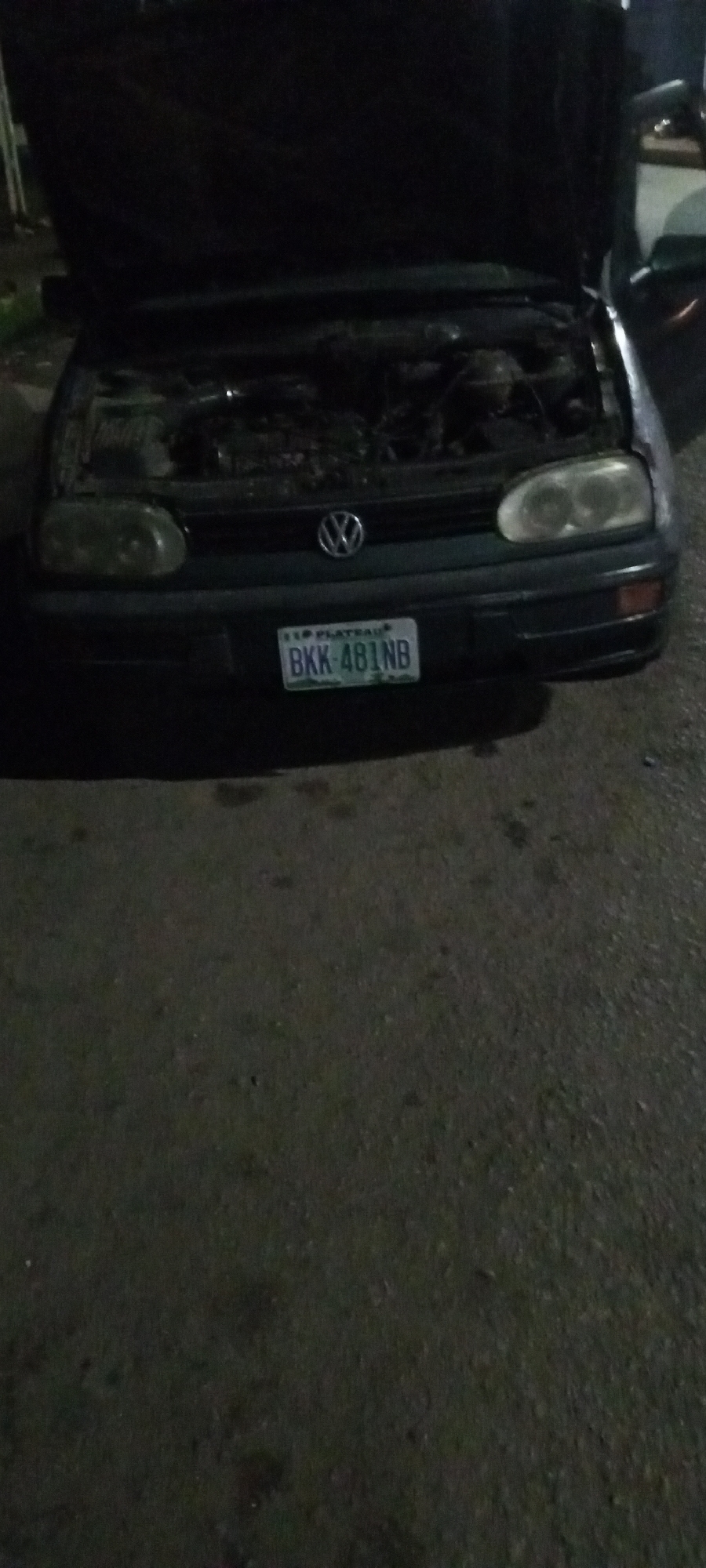 An open-hooded, dark-colored Volkswagen Golf is parked on an asphalt surface at night. The car's license plate reads "BKK 481NB" and is located below the car's emblem. The scene is dimly lit, suggesting it is nighttime. There are no people visible in the image. The environment appears to be an outdoor area, possibly a street or parking lot, with some scattered debris on the asphalt.