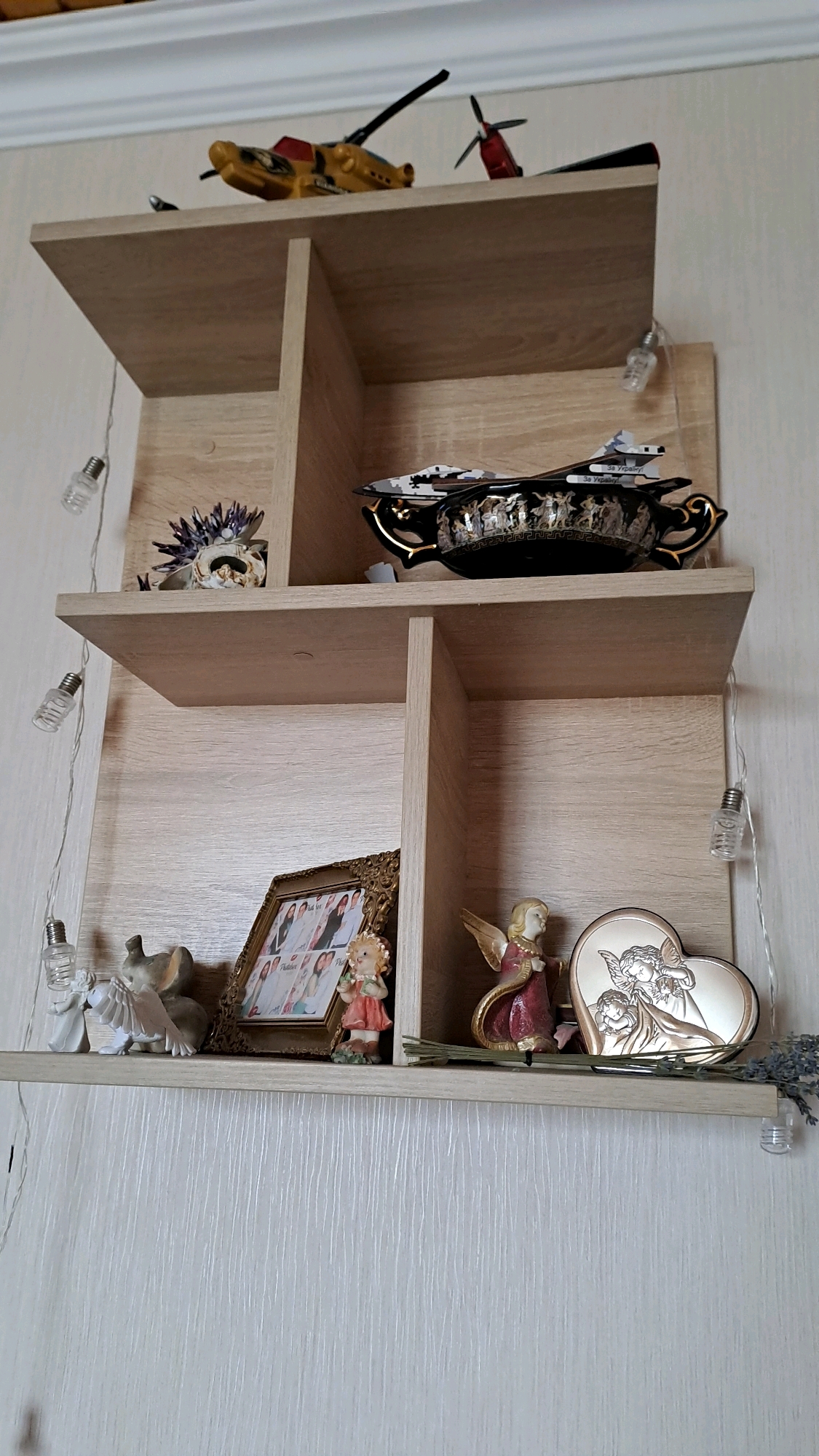 The image shows a wooden shelf unit with several decorative items. On the top shelf, there are toy military vehicles, including a yellow tank and a red airplane. Below that, on the left side, is a small sculpture of a face with purple flowers. On the right side of the second shelf, there is a black decorative bowl with gold accents and a scene of figures. The third shelf, on the left, contains a framed photo of several people, a small figurine of a girl in a pink dress, and a figurine of an elephant. On the right side of the third shelf, there is a figurine of an angel in a red dress and a heart-shaped object with an angel motif. There are also strings of small lights draped around the shelves. The background appears to be a textured wall. The image is likely taken indoors.
